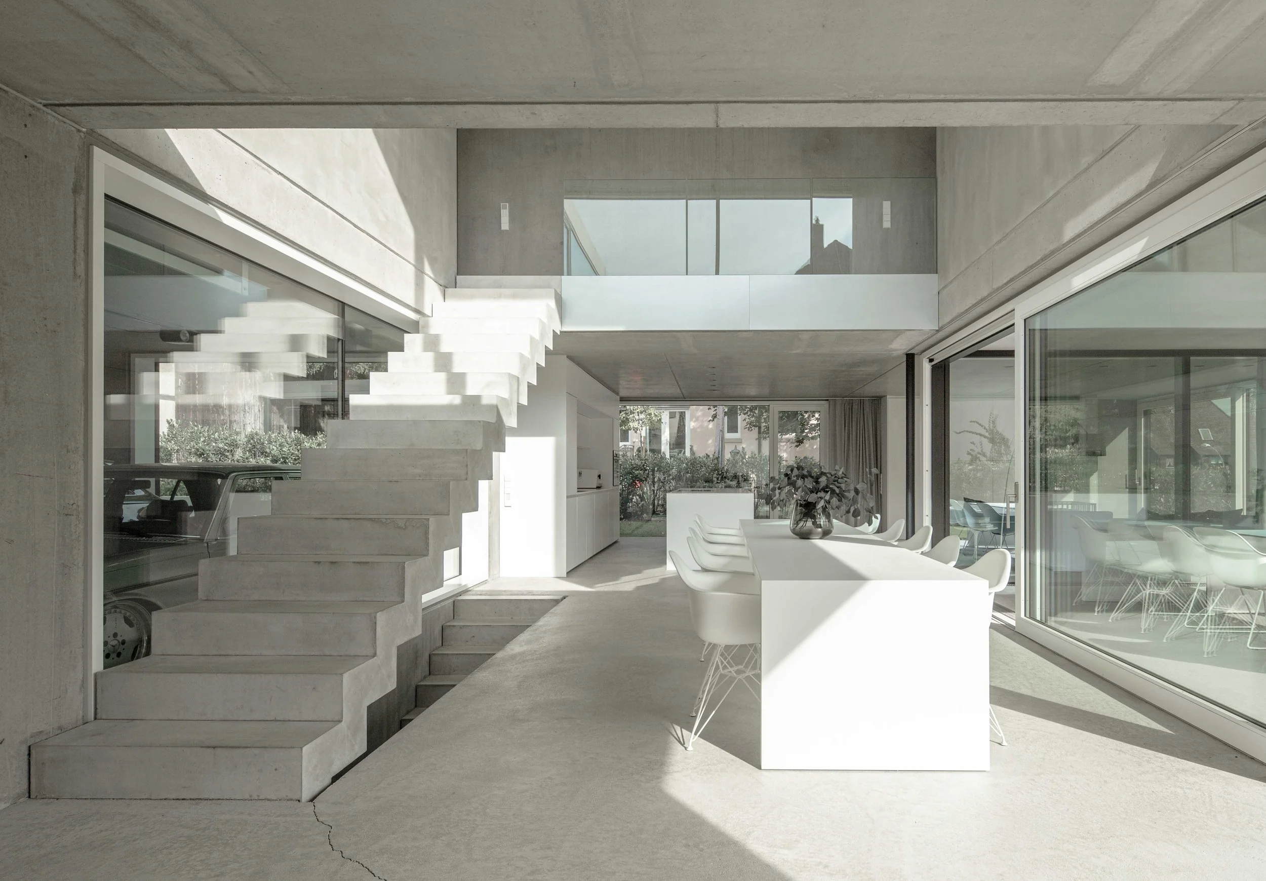 haus vO - Innenansicht eines modernen, minimalistischen Hauses mit Betonwänden, Treppe, großen Glasfenstern und einem Essbereich mit weißen Stühlen und Tisch.