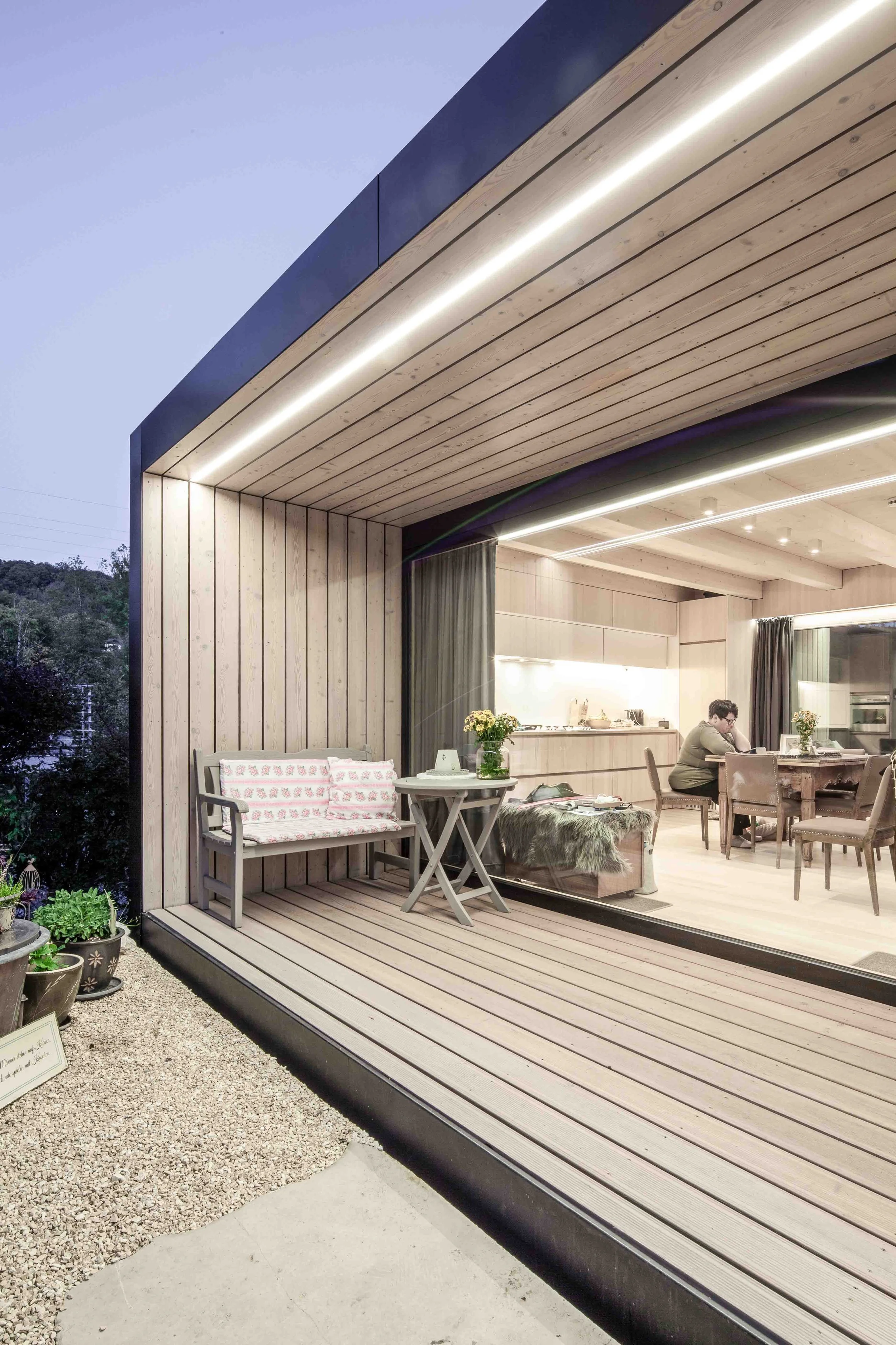 haus GRO - Gemütlicher Balkon mit Sitzgelegenheit, Holzboden und Blick ins Innere eines modernen Wohnzimmers mit Esstisch und Küchenbereich. Draußen Pflanzen auf Kieselsteinen.