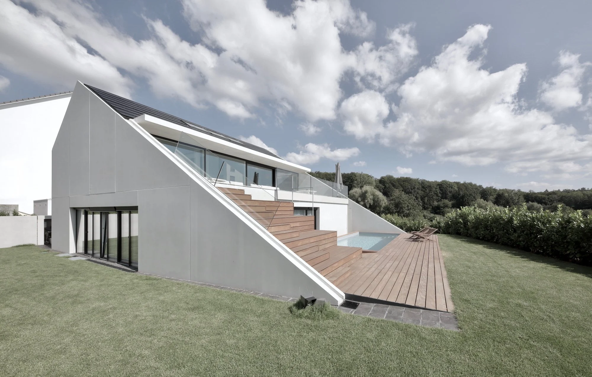 haus CC - Modernes, weißes Haus mit großen Glasfenstern, Holzterrasse, Pool und Garten, umgeben von Bäumen unter einem bewölkten Himmel.