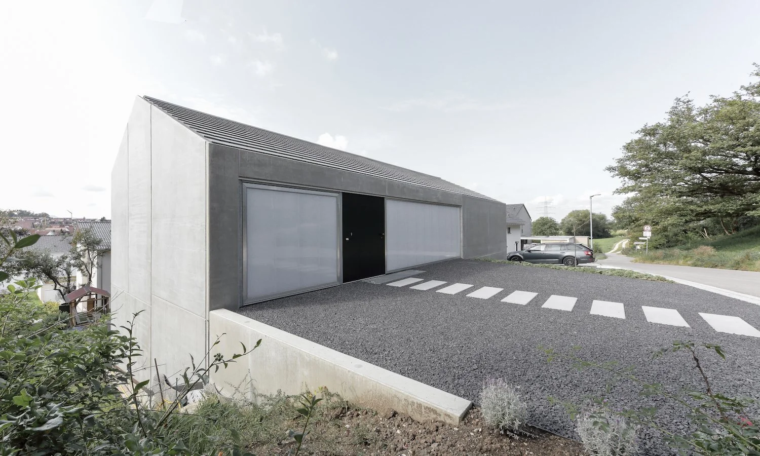 haus WÄ - Moderner grauer Holzschuppen mit großen, weißen Türen, umgeben von Bäumen und einem Parkplatz mit Auto, im Hintergrund eine Straße und Wohnhäuser, sonniger Tag.