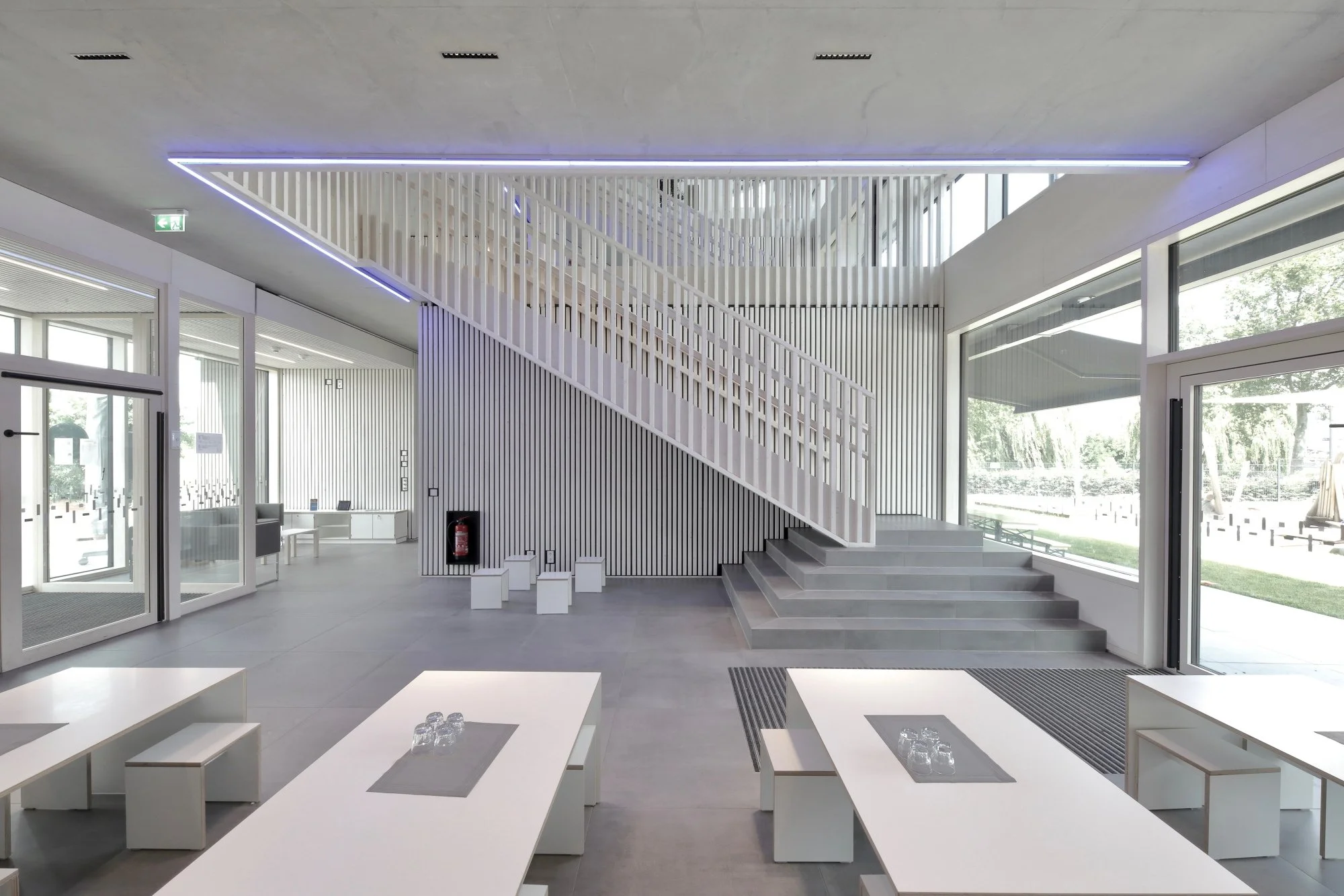 kinderhaus BUGA - Innenraum eines modernen Gebäudes mit großen Fenstern, weißen Möbeln, Treppe aus Holz und Lichtinstallationen an der Decke.