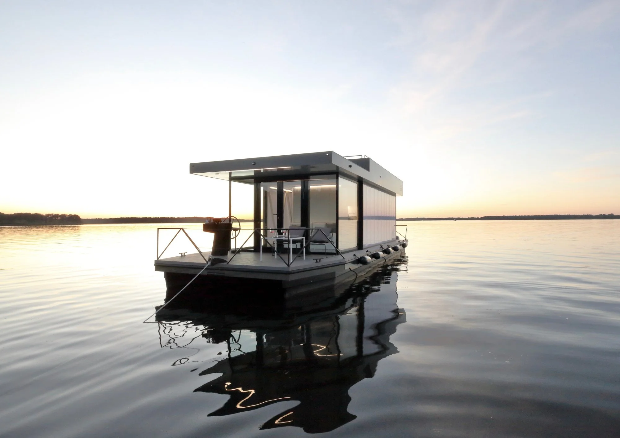 Ein modernes Hausboot liegt auf einem ruhigen See, während die Sonne untergeht, mit einem weichen, goldenen Licht und einem klaren Himmel im Hintergrund.