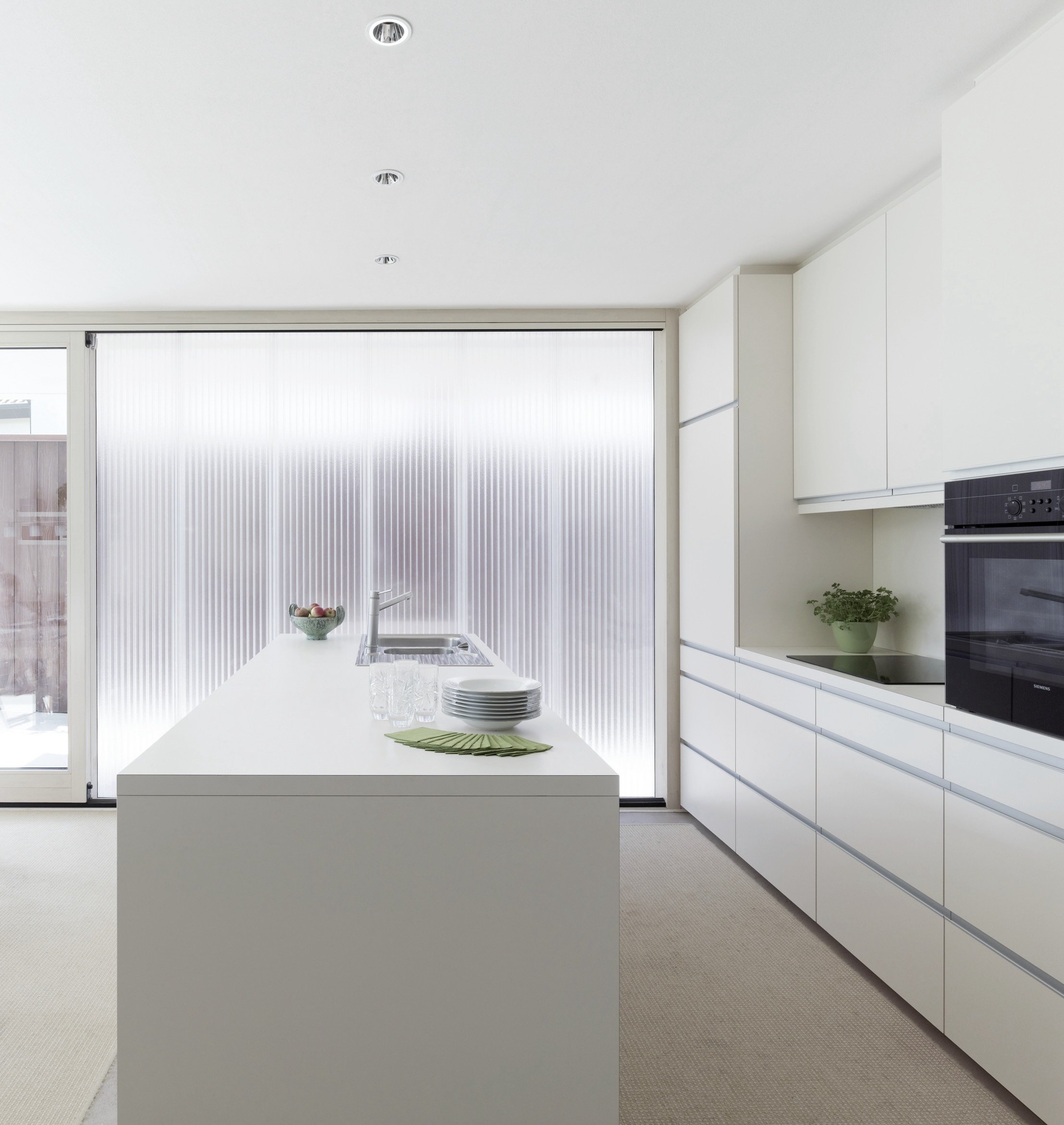 haus - B_Helle moderne weiße Küche mit Kücheninsel, Spüle, Geschirr, Obstschale, Einbauschränken und Backofen.