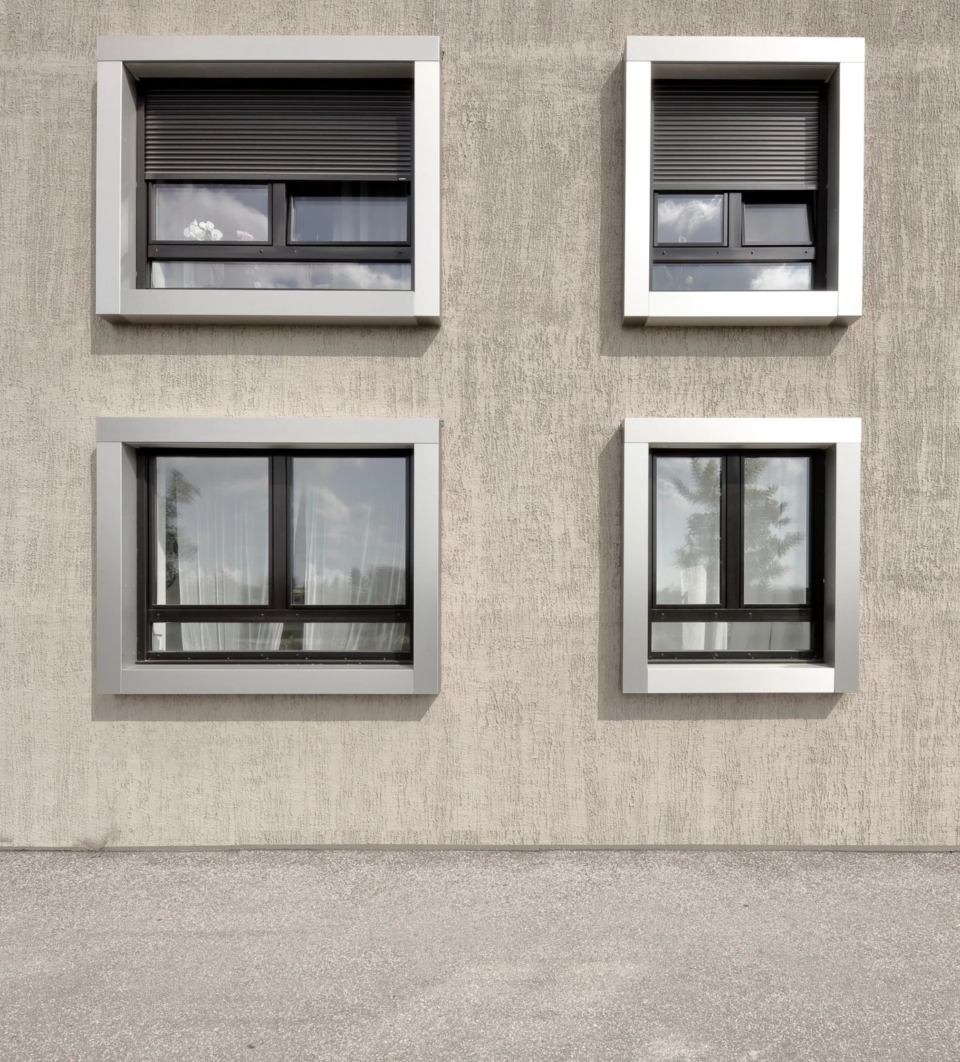wohnhaus BUGA - Vier quadratische Fenster in einer beige Wand, mit weißen Rahmen und schwarzen Jalousien, zwei Fenster im oberen Bereich, zwei im unteren Bereich, mit Reflexionen des Himmels und Bäumen sichtbar.