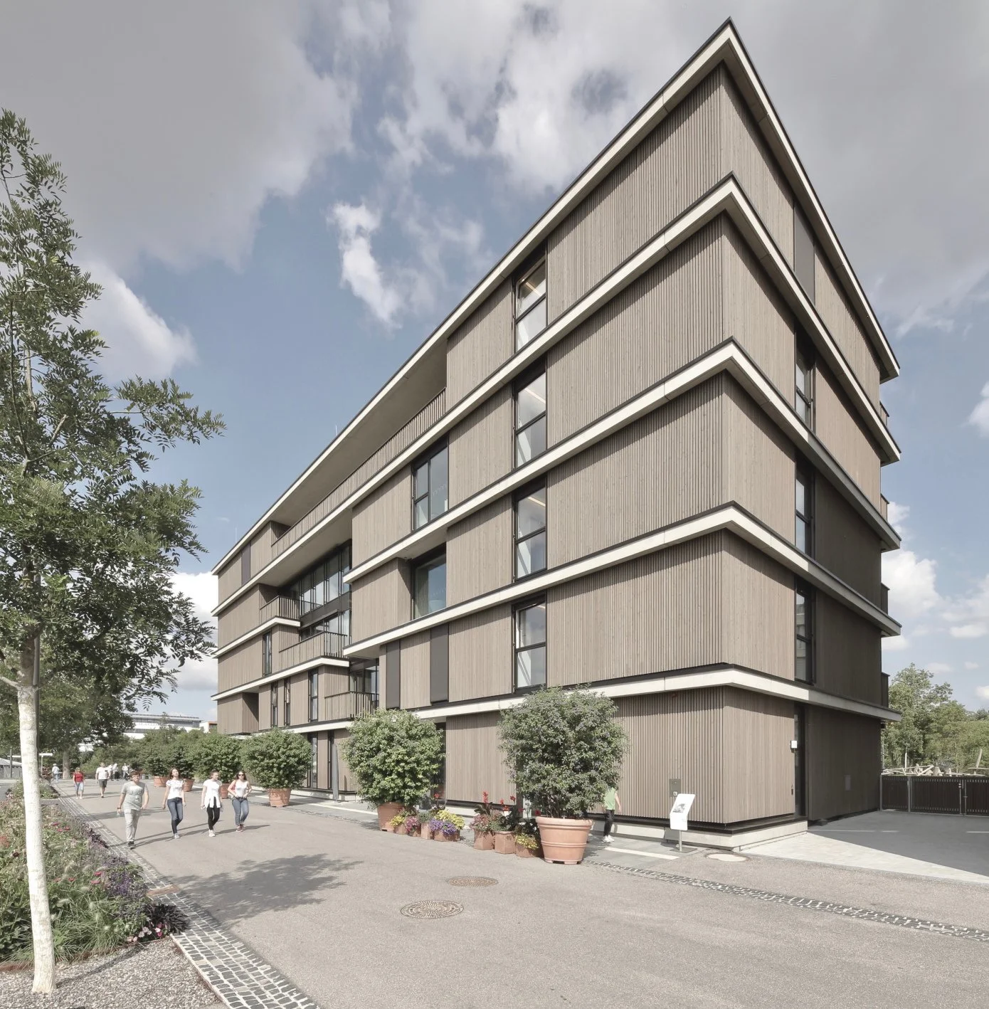 kinderhaus BUGA - Mehrstöckiges modernes Wohngebäude mit Holzverkleidung, umgeben von Bäumen und Fußgängern auf einem Bürgersteig.