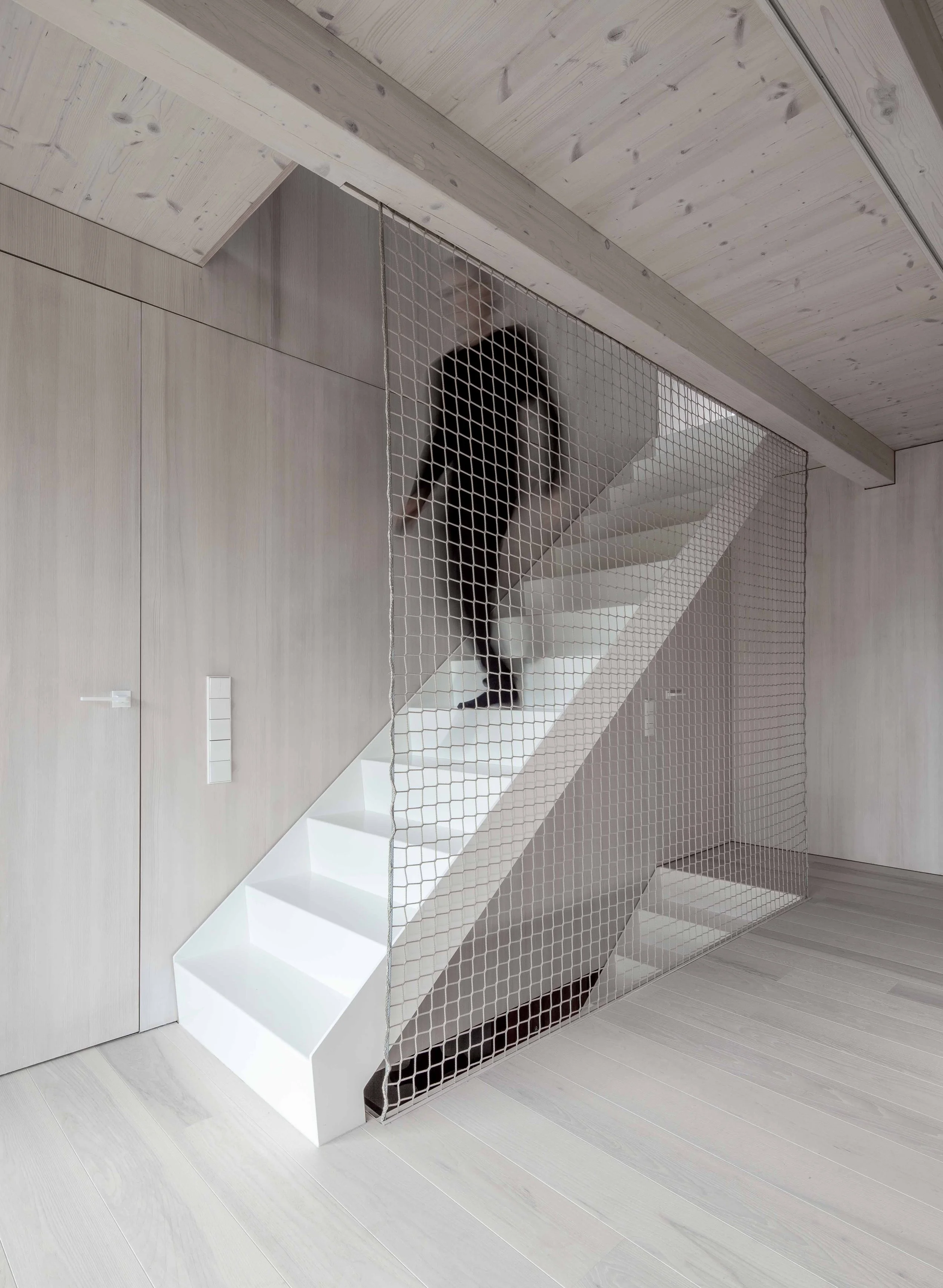 haus GRO - Weißer moderner Kamin mit einer weißen Sitzbank daneben, Raum mit hellen Holzwänden und Boden, Sicht auf eine Treppe mit dunklen Stufen durch eine Gittertrennwand.