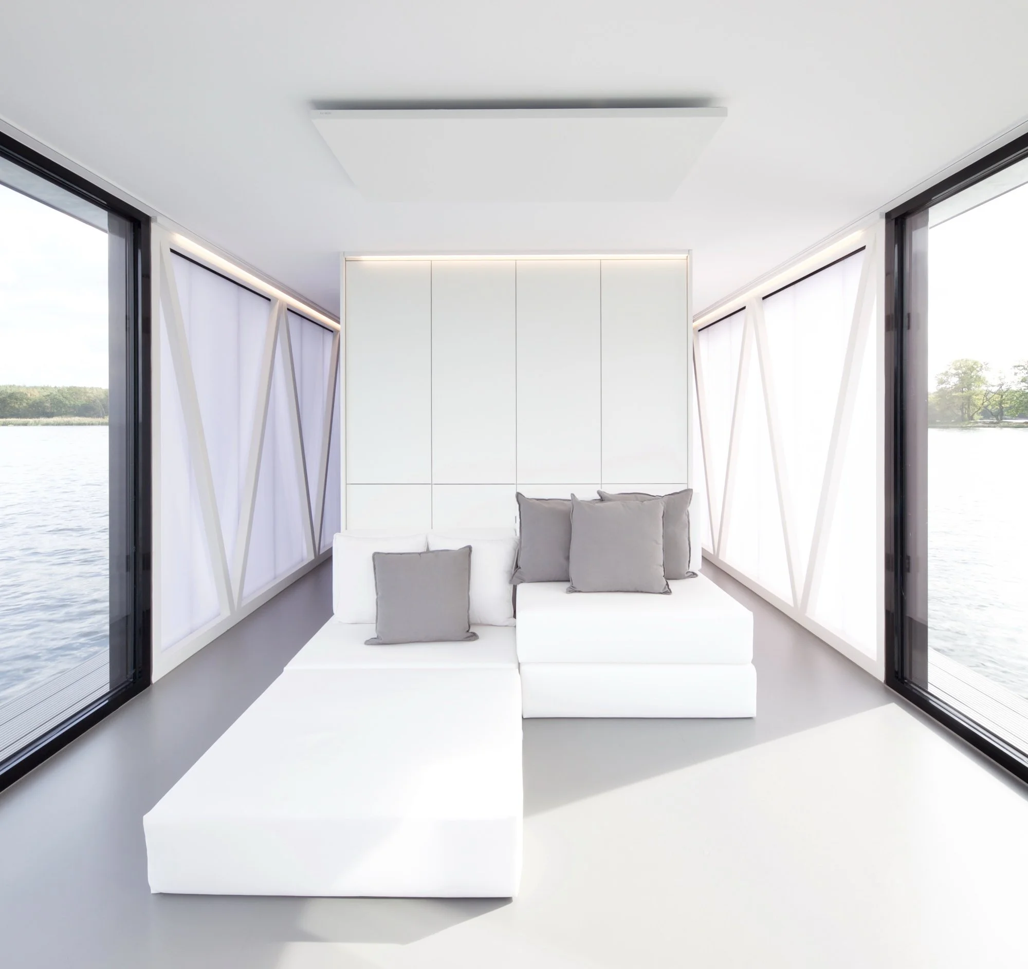 Moderner Raum mit großen Fenstern, weißem Sofa, mehreren Kissen und Blick auf einen Fluss im Hintergrund.