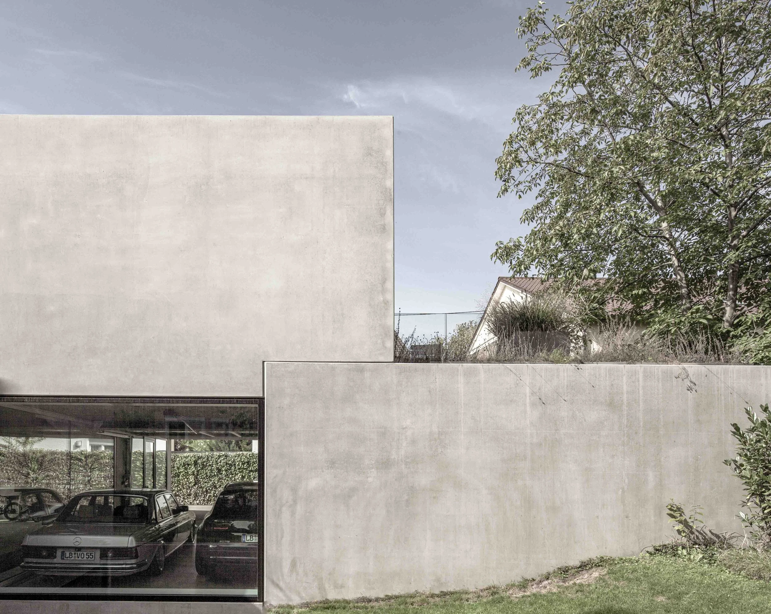 haus vO - Ansicht eines modernen Gebäudes mit einer weißen Fassade und großen Glasflächen im Erdgeschoss. Im Vordergrund befinden sich Autos in einer Tiefgarage. Auf der rechten Seite sieht man Bäume und einen Teil eines Blumensпрücks.