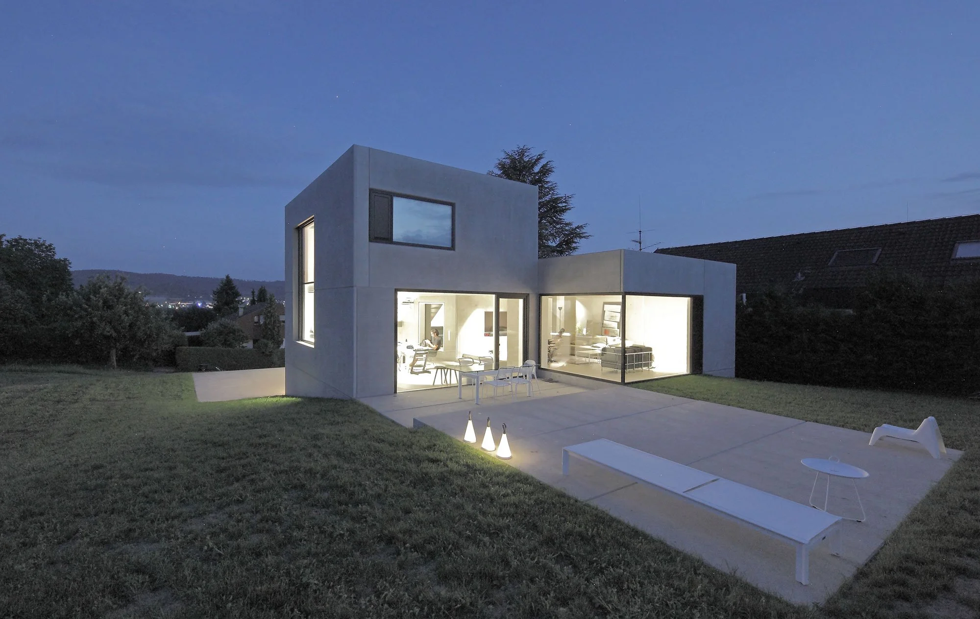 haus SG - Moderne weiße Villa bei Nacht mit beleuchteten Innenräumen, Gartenmöbeln und umgebender Natur.