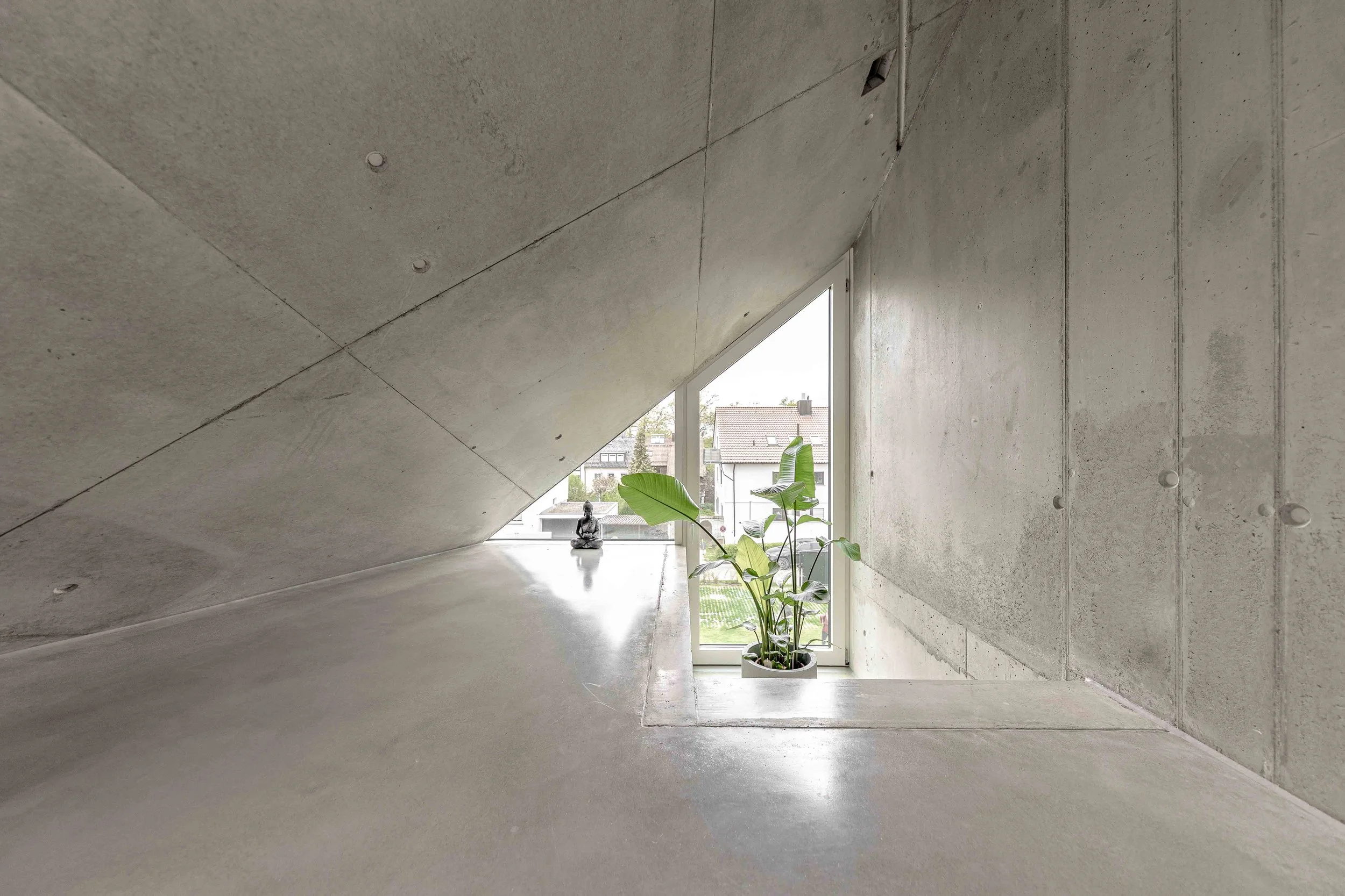 Innenraum eines modernen Gebäudes mit rohem Beton, großer Fensterfront und Pflanzen, eine sitzende Figur bei der Fensteröffnung.