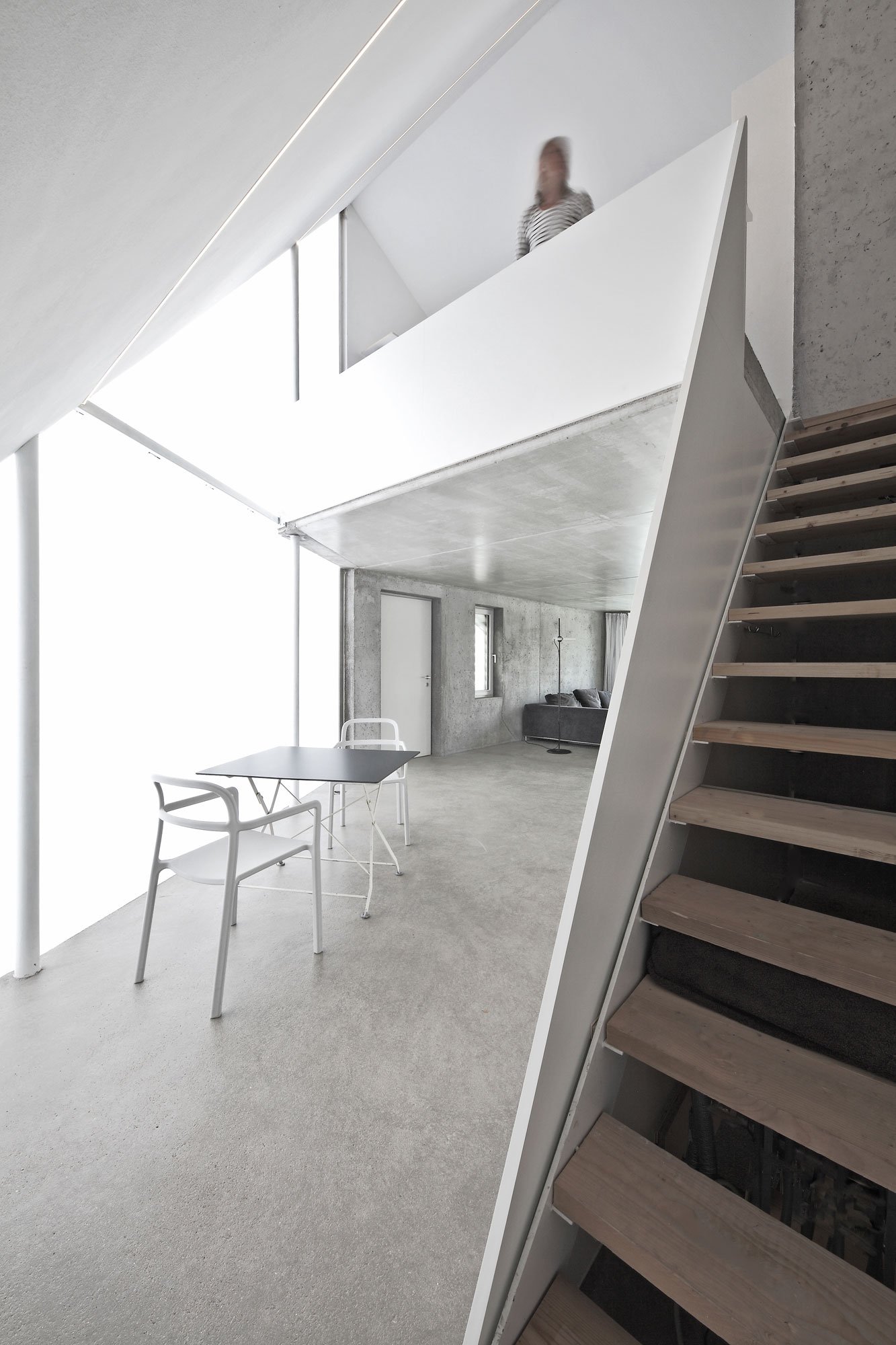haus SO - Innenraum eines modernen, minimalistischen Wohnraums mit Treppe, Glasfronten, einem hohen Fenster, einem Tisch mit zwei Stühlen, einem Sofa und einem Stehlampe, alles in neutralen Farben.