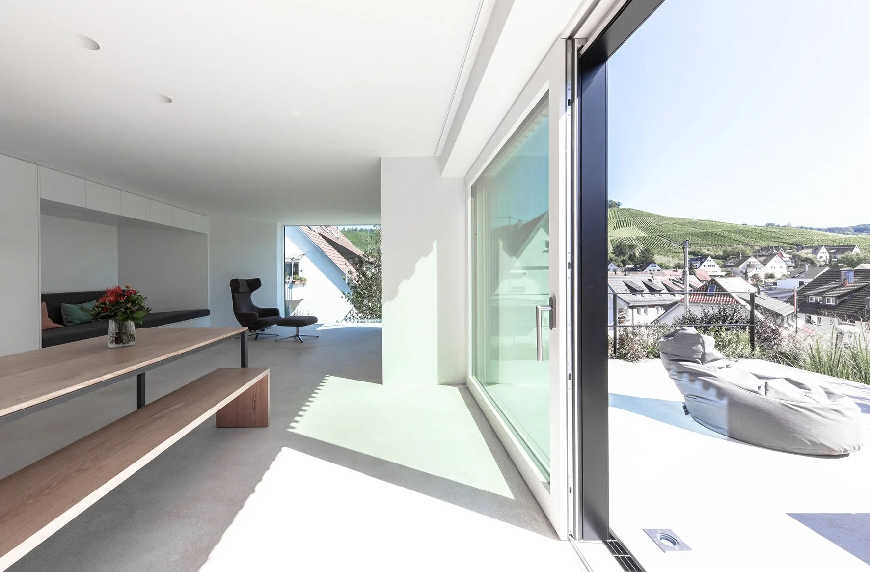 haus RU - Modernes Wohnzimmer mit großen Fenstern, Blick auf ein Dorf und Weinberge im Hinterland, minimalistische Einrichtung, bequemer Sessel, Holzbank, Blumenstrauß. Außenbereich mit Liege auf Terrasse.