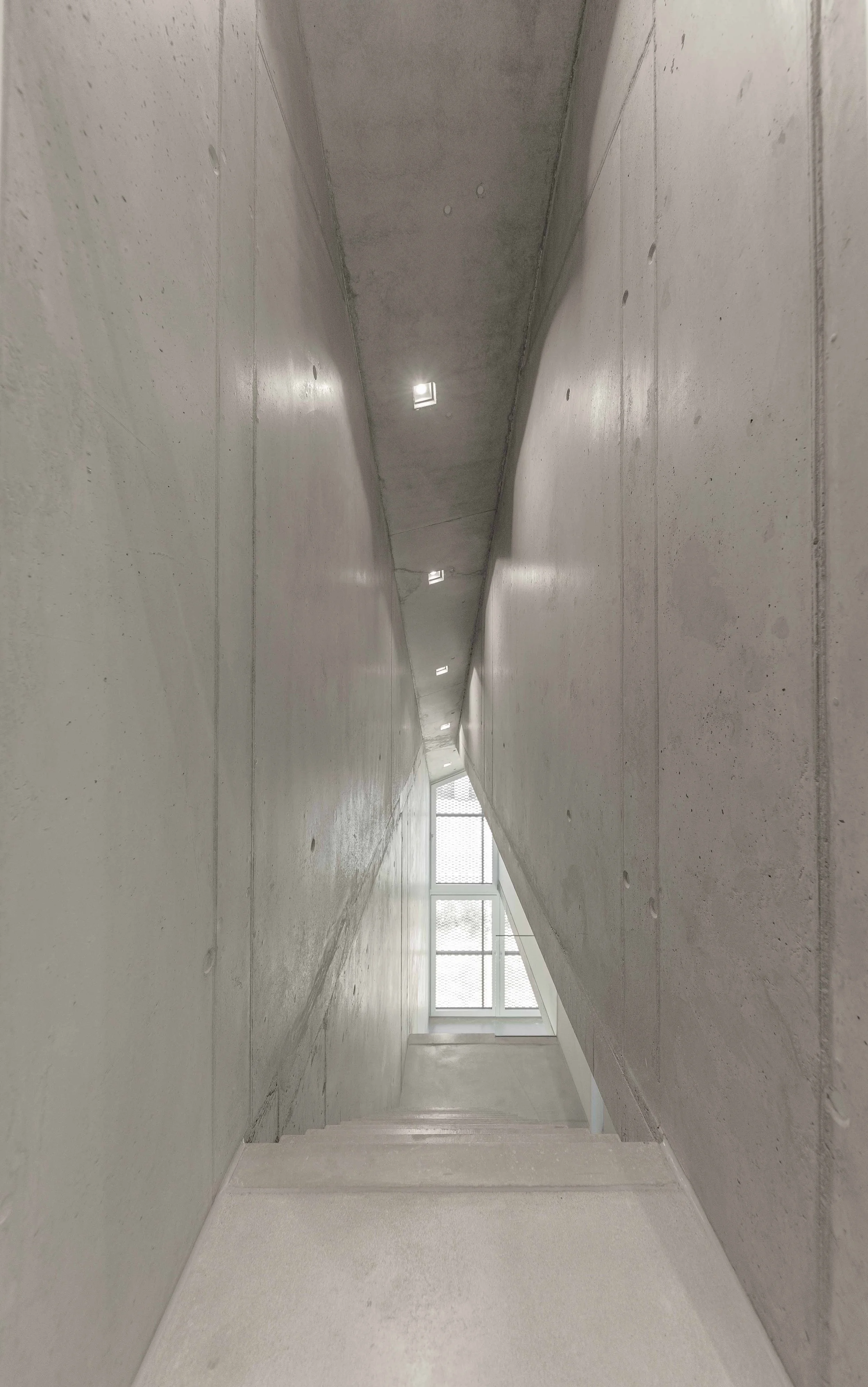 Ein heller, moderner Tunnel mit glatten Betonwänden und Boden, mit einigen Deckenlichtern und großen Fenstern am Ende, die Tageslicht hereinlassen.