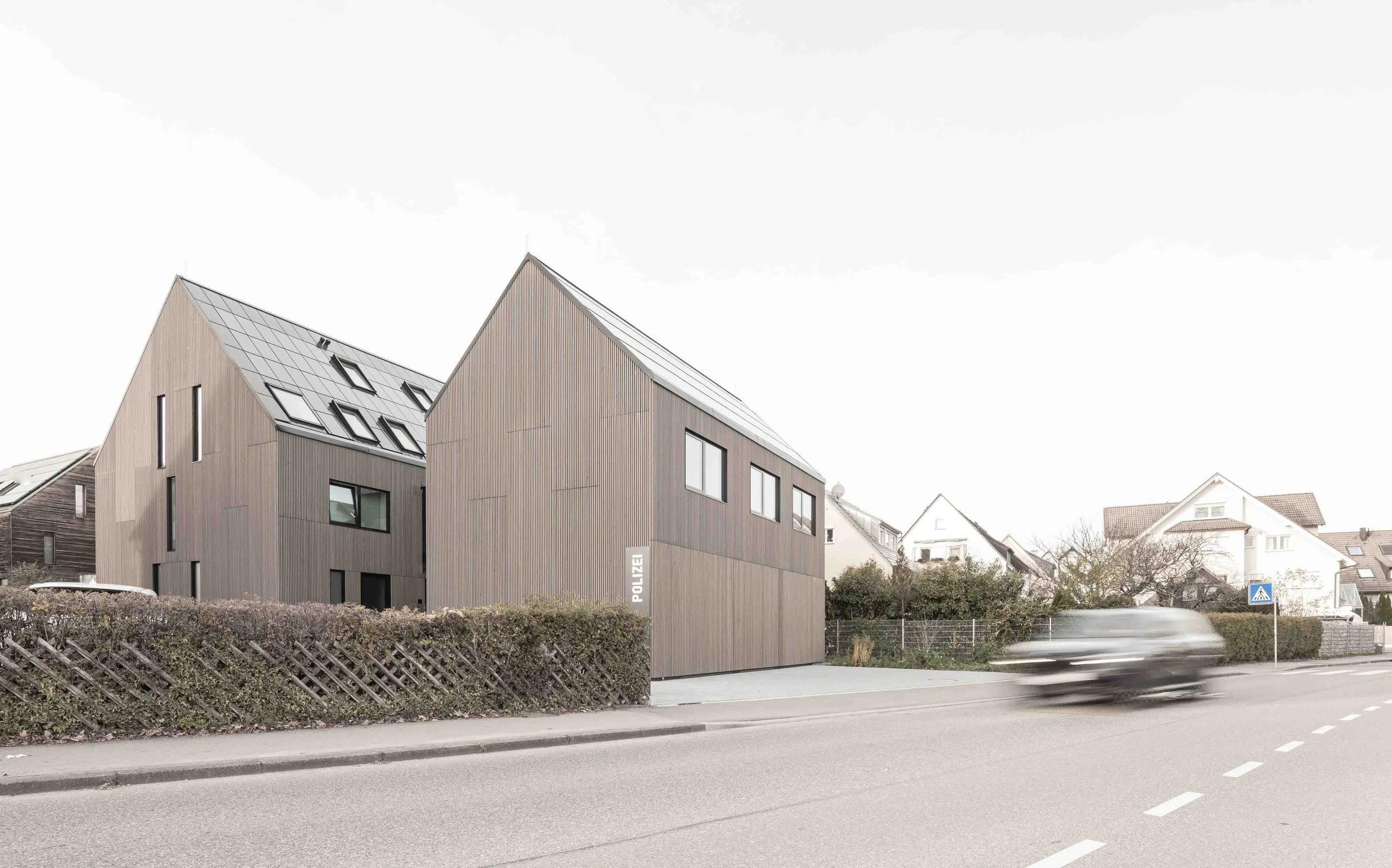 polizeiposten LE - Moderne, zweistöckige Polizeistation mit Holzfassade und mehreren Dachfenstern, angrenzender Weg und überfahrendes Fahrzeug, einige Wohnhäuser im Hintergrund, bei Tageslicht.