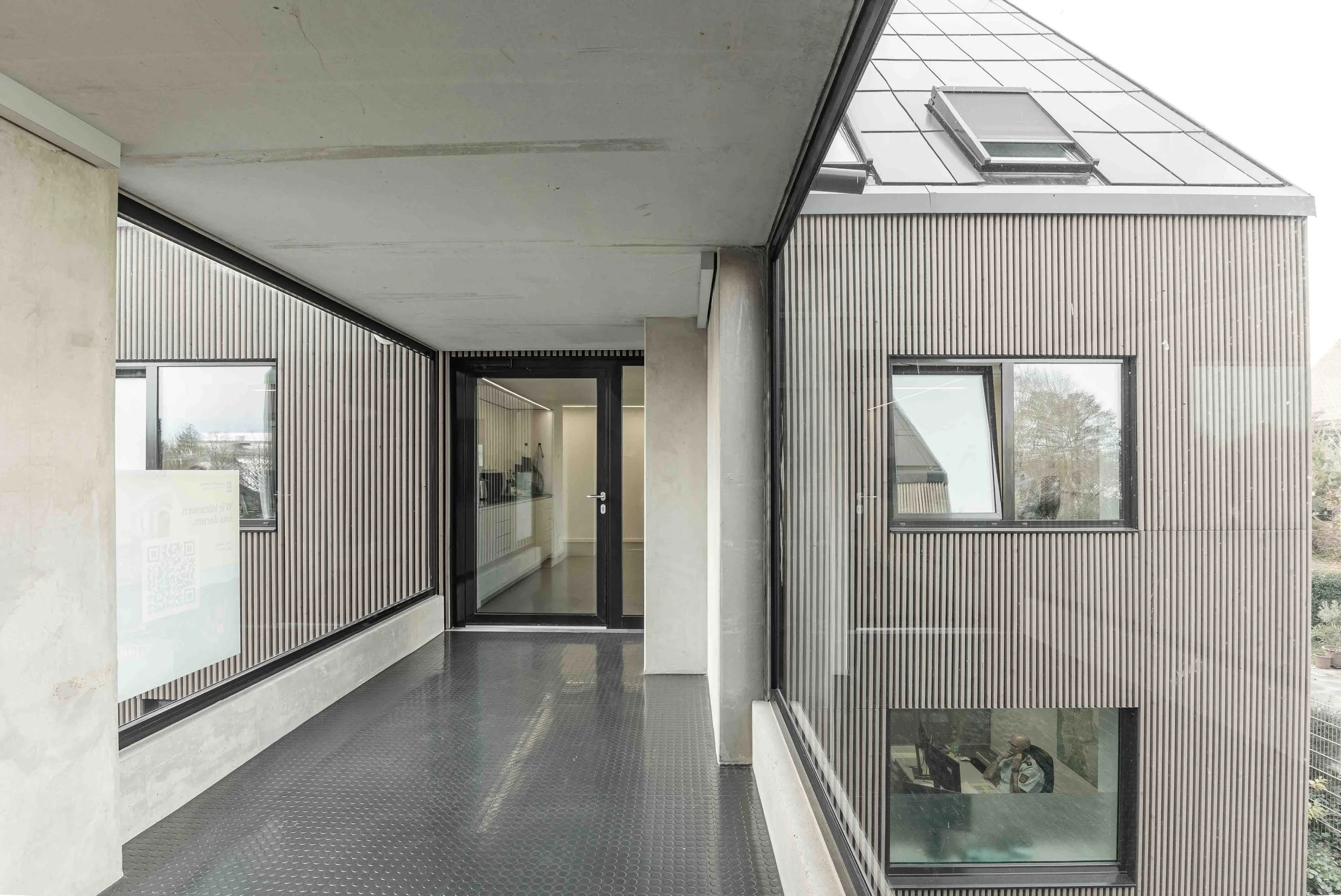 polizeiposten LE - Moderner Eingangsbereich eines Gebäudes mit großen Fenstern, gepflastertem Boden und minimalistischer Architektur aus Beton und Holz.