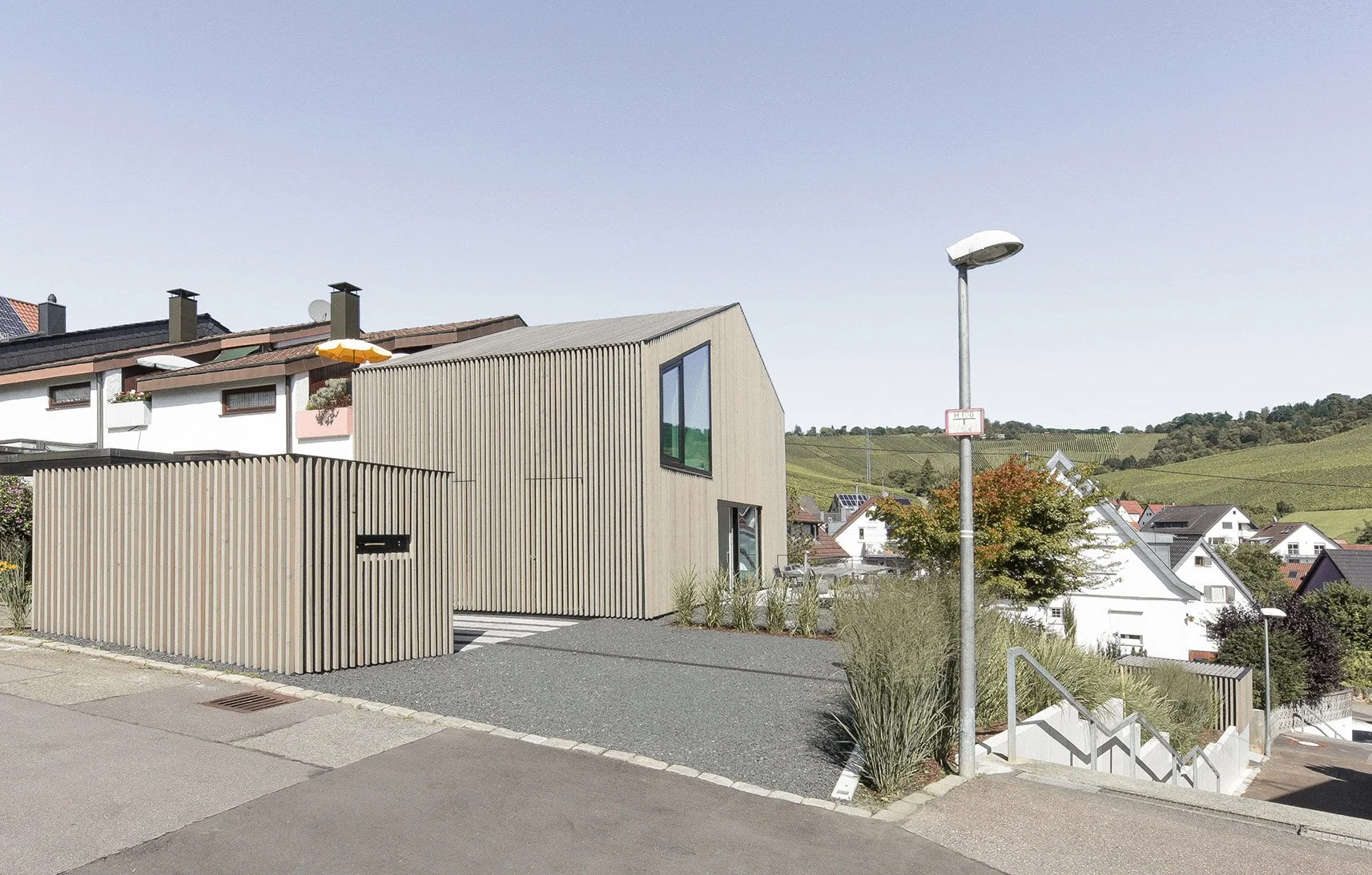 haus RU - Modernes zweistöckiges Haus mit vertikalen Holzpaneelen, großen Fenstern und gartenartige Umgebung in einem ländlichen Gebiet mit Hügeln im Hintergrund.