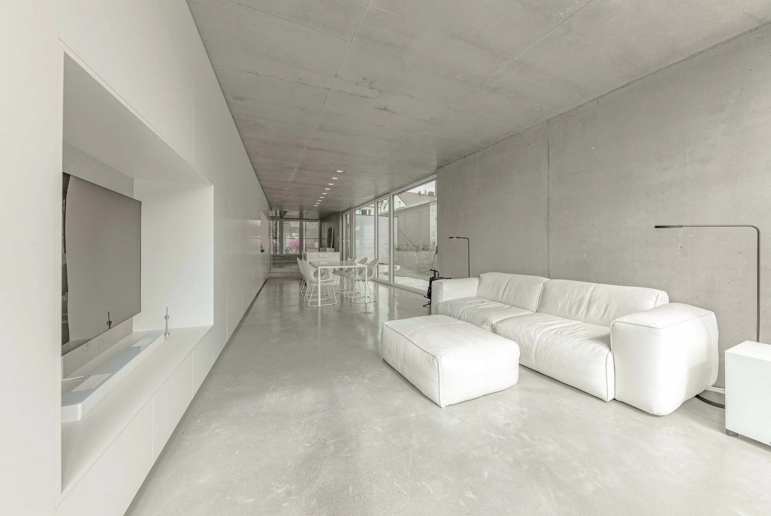 Modernes Wohnzimmer mit weißen Möbeln, großen Fenstern und einer Betonwanddecke.
