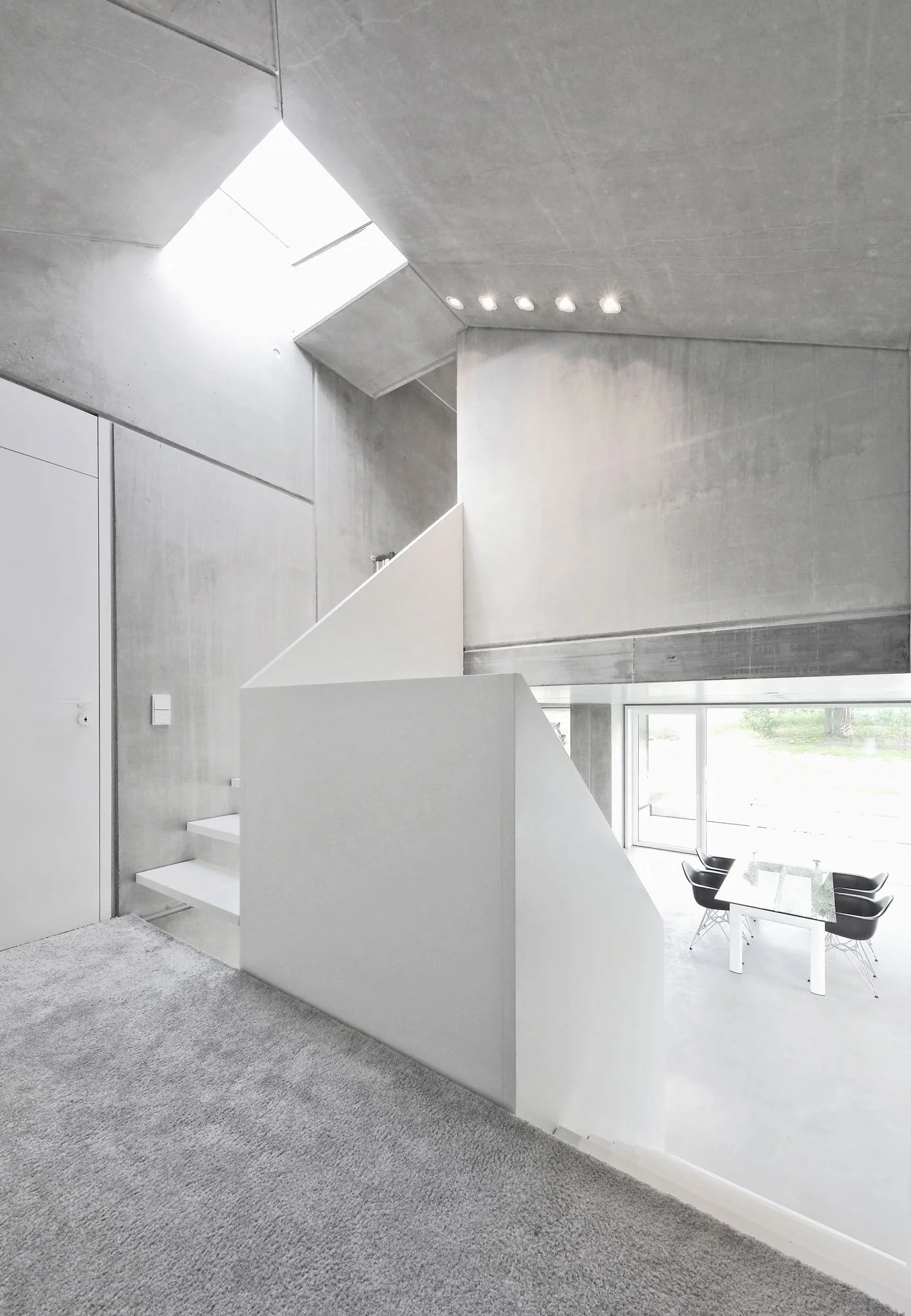 haus SG - Moderne, helle Wohnung mit offener Raumgestaltung, grauem Teppichboden, weißen Wänden und Blick auf eine Küche und einen Essbereich mit schwarzem Stuhl und Glastisch.