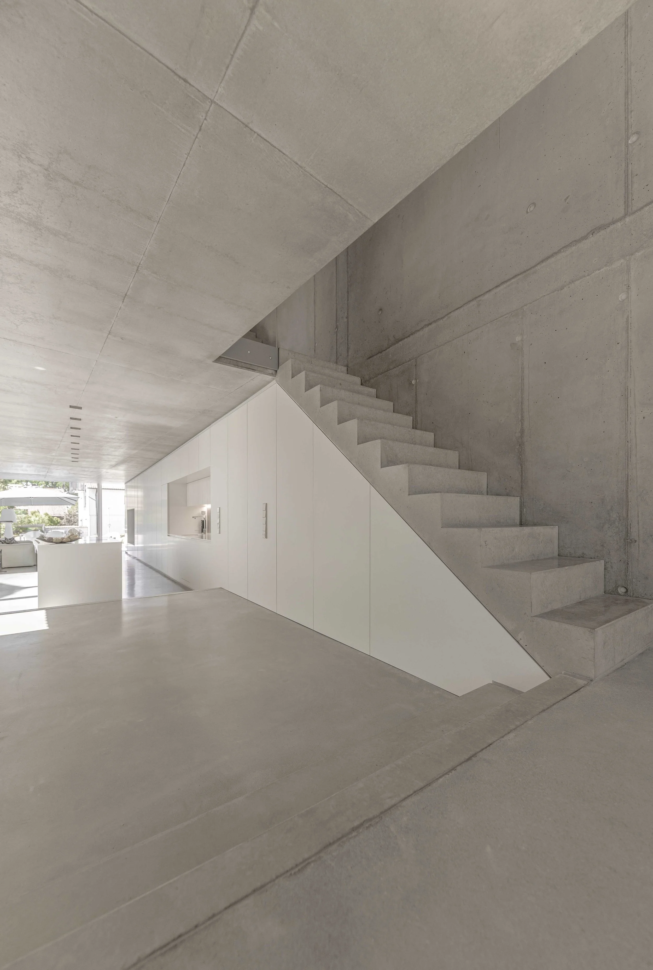 Innenraum eines modernen Gebäudes mit einer grauen Betonwand, Treppe und weißen Wänden, durch Fenster ist ein Außenbereich sichtbar.