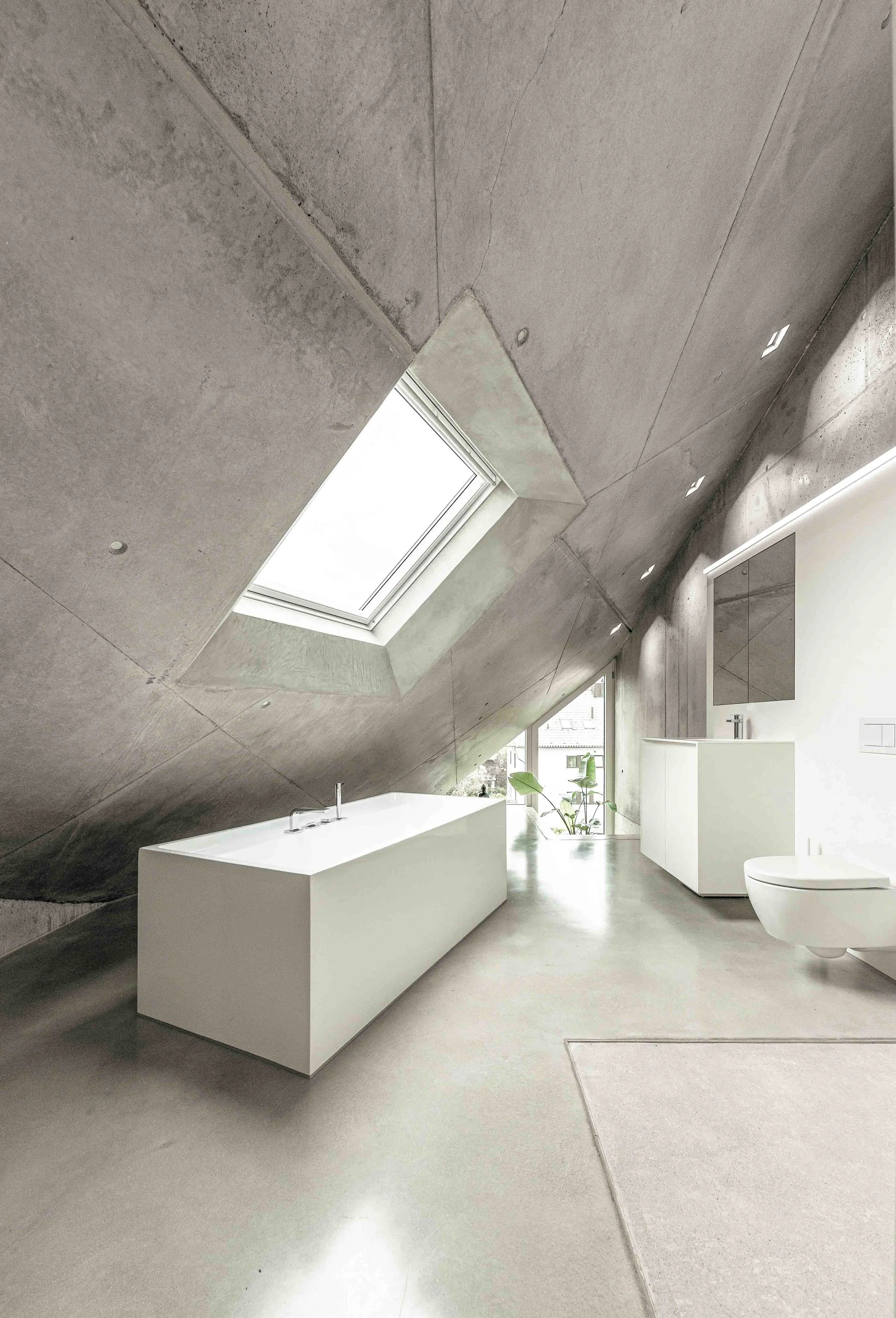 Ein modernes, minimalistisch gestaltetes Badezimmer mit einer weißen Badewanne, einem weißen Waschbecken, einem spülkasten, einem Fenster in der Dachschräge, einer Pflanze und weißen Möbeln, in einem Raum mit Betonwänden und weitem, hellen Raum.