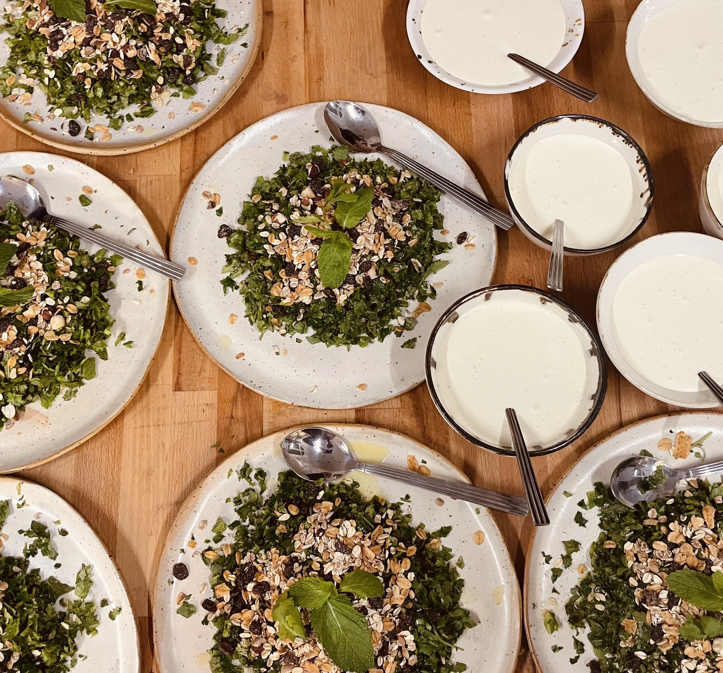 Billede af flere tallerkener med grøn salat drysset med nødder og tørret frugt, og flere glas med hvid væske, som kan være yoghurt eller sauce, placeret på en træbord.