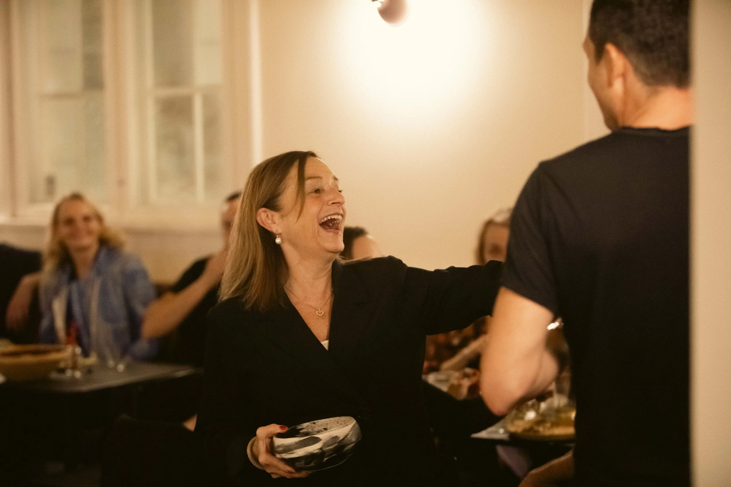 Kvinde og mand, der smiler og taler, mens de deler et øjeblik ved en social sammenkomst, med flere mennesker i baggrunden, der sidder ved et bord.