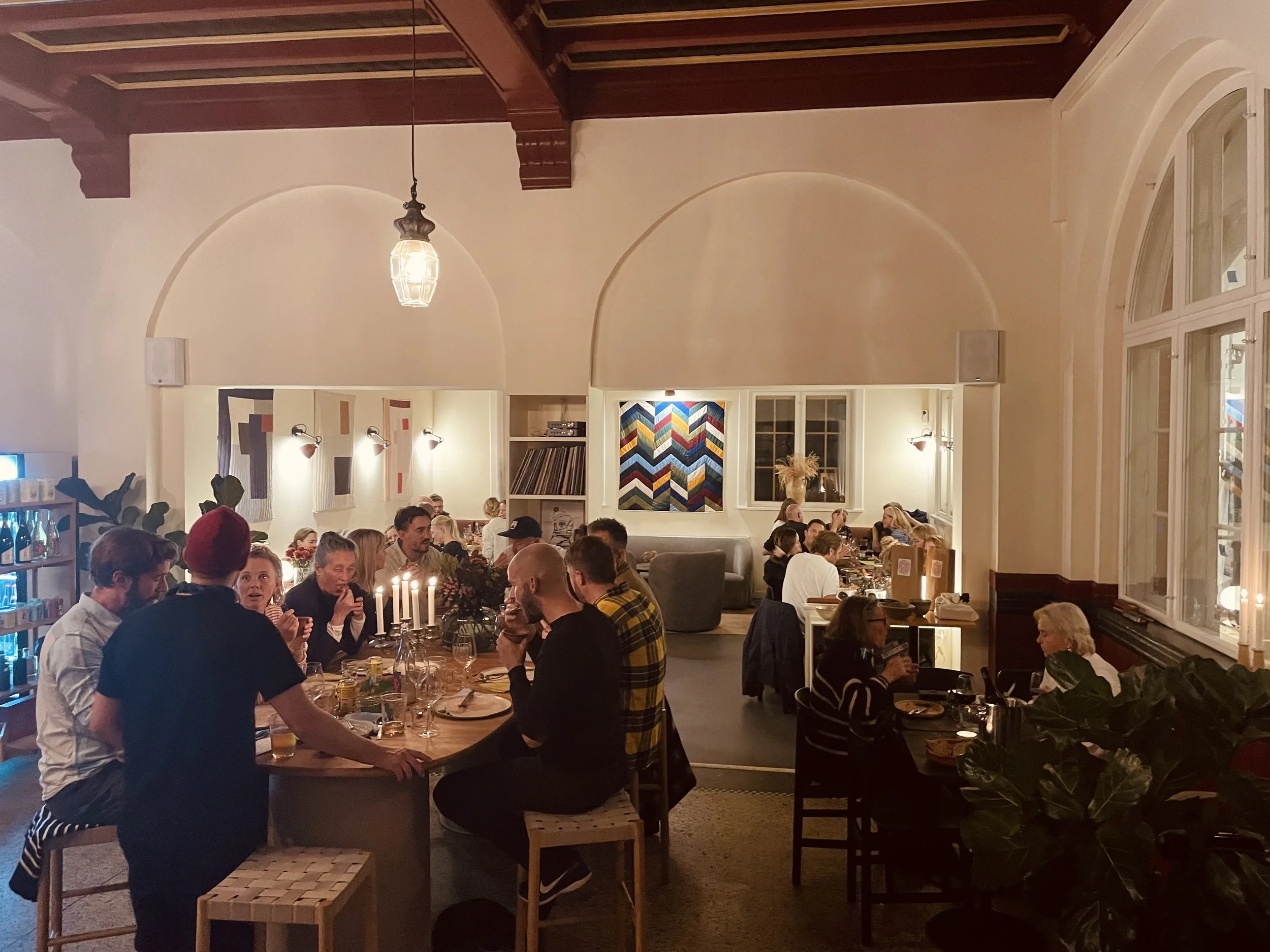 Indendørs restaurant med flere mennesker, der spiser og snakker, varme belysning, store vinduer og moderne kunst på væggen.