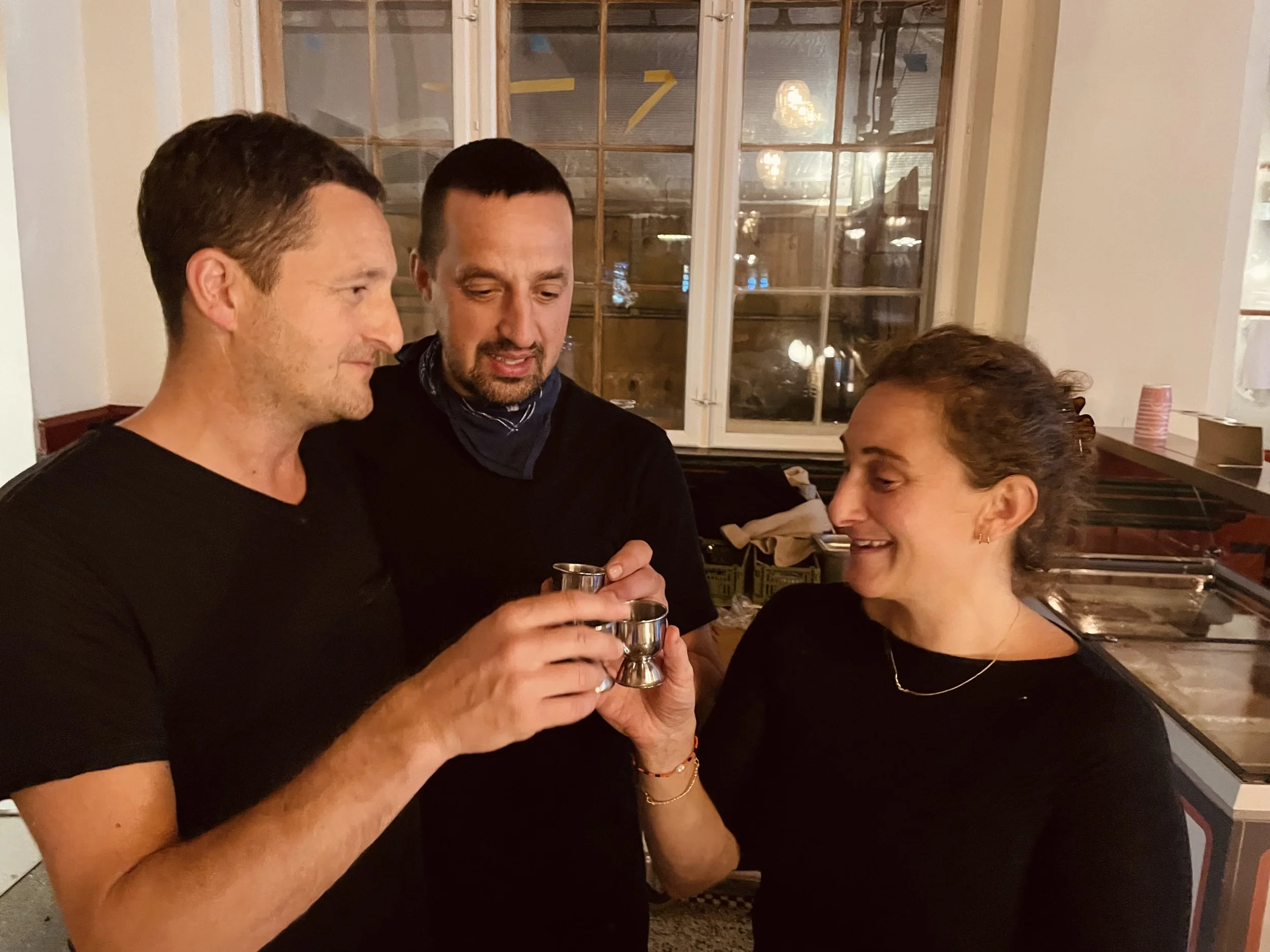 Tre personer skåler med små metalshots i et indendørs miljø med store vinduer i baggrunden.