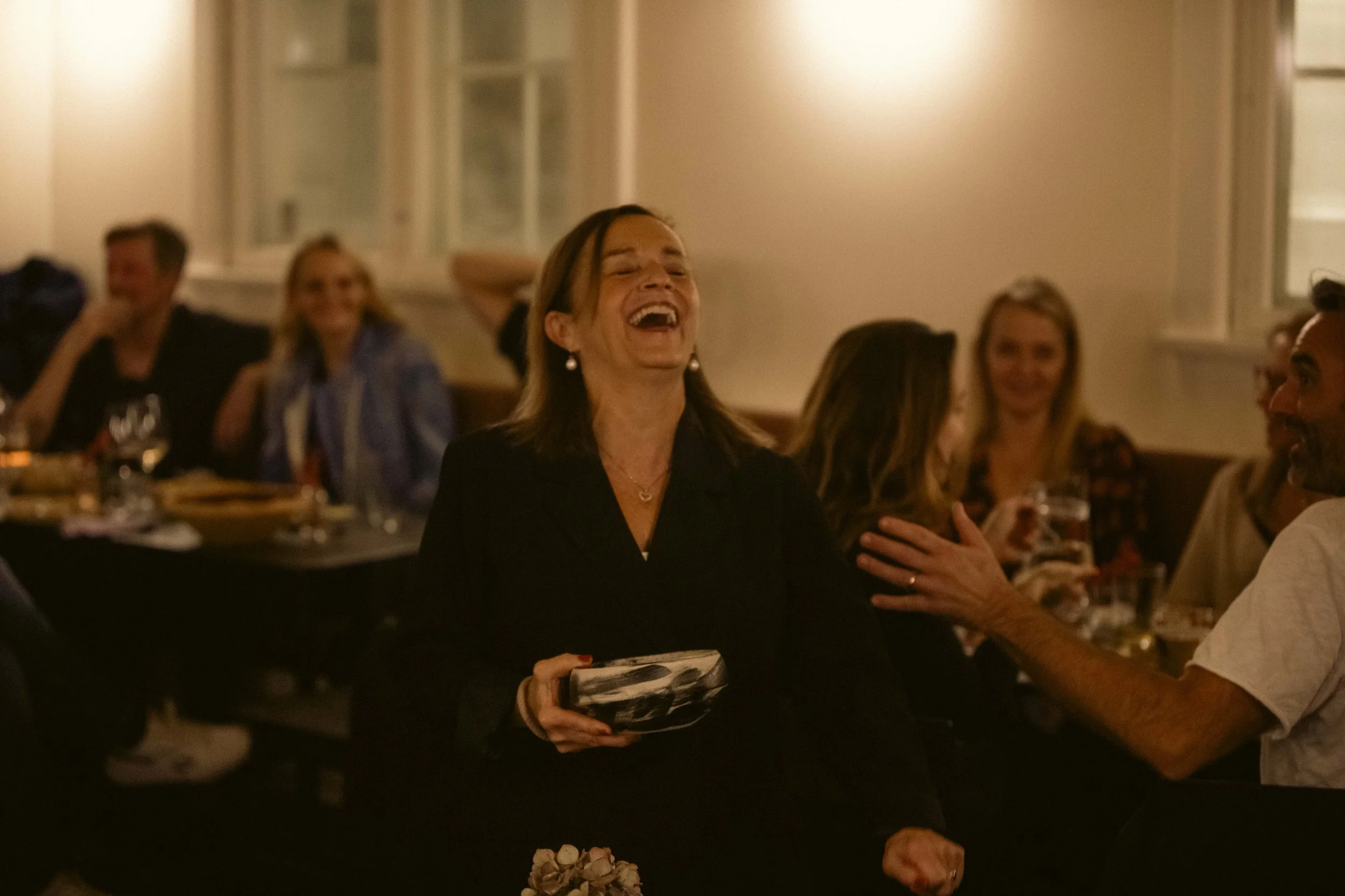 En kvinde ler og holder en tallerken, mens hun sidder ved et bord omgivet af andre mennesker i en hyggelig indendørs fest eller middag.