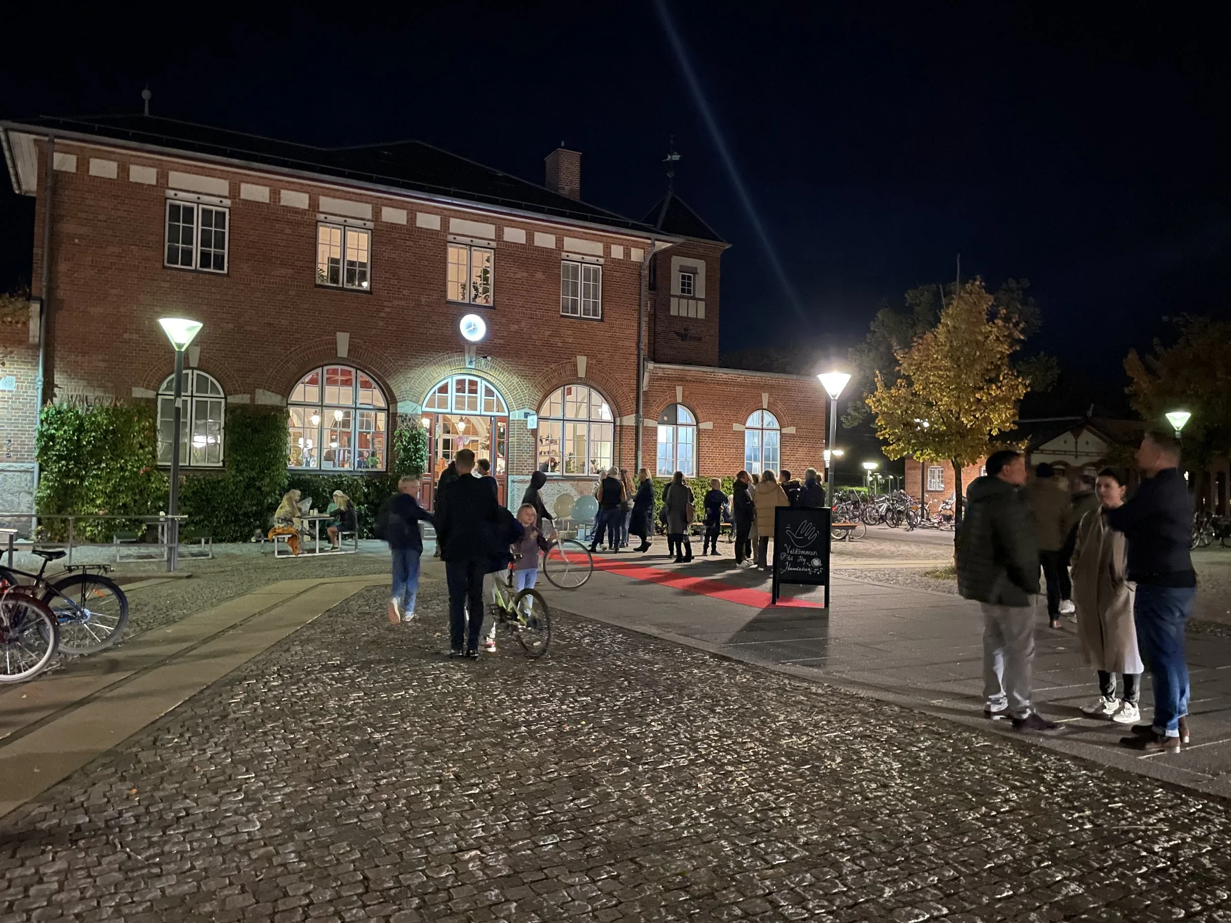 En gruppe mennesker står og snakker udenfor en røde murstensbygning om aftenen. Der er cykler parkeret, og nogle personer sidder på bænke. Gadebelysning lyser området op, og der er tilhørende træer med efterårsblade.