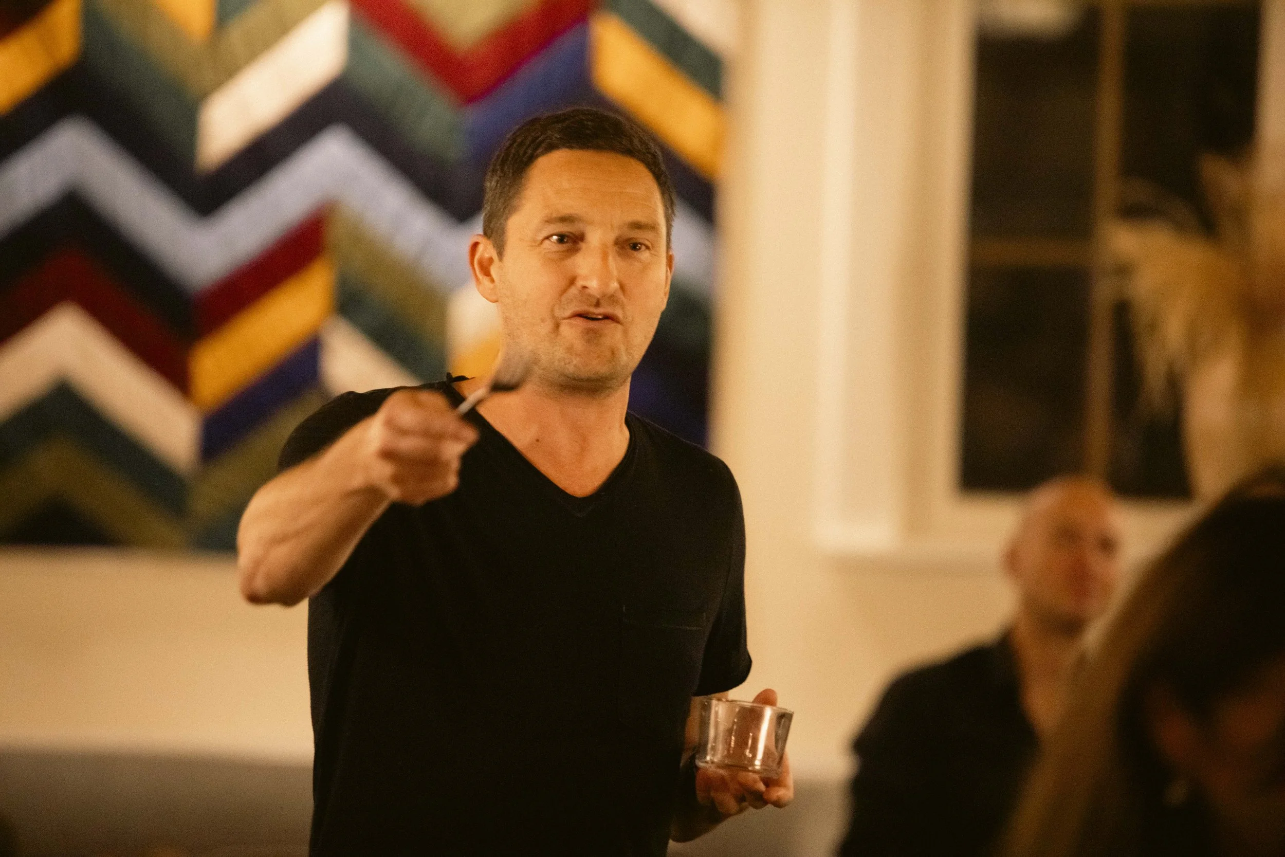 En mand i sort t-shirt holder en drink og peger mod kameraet i en lejlighed med farverigt vægkunst og andre personer i baggrunden.