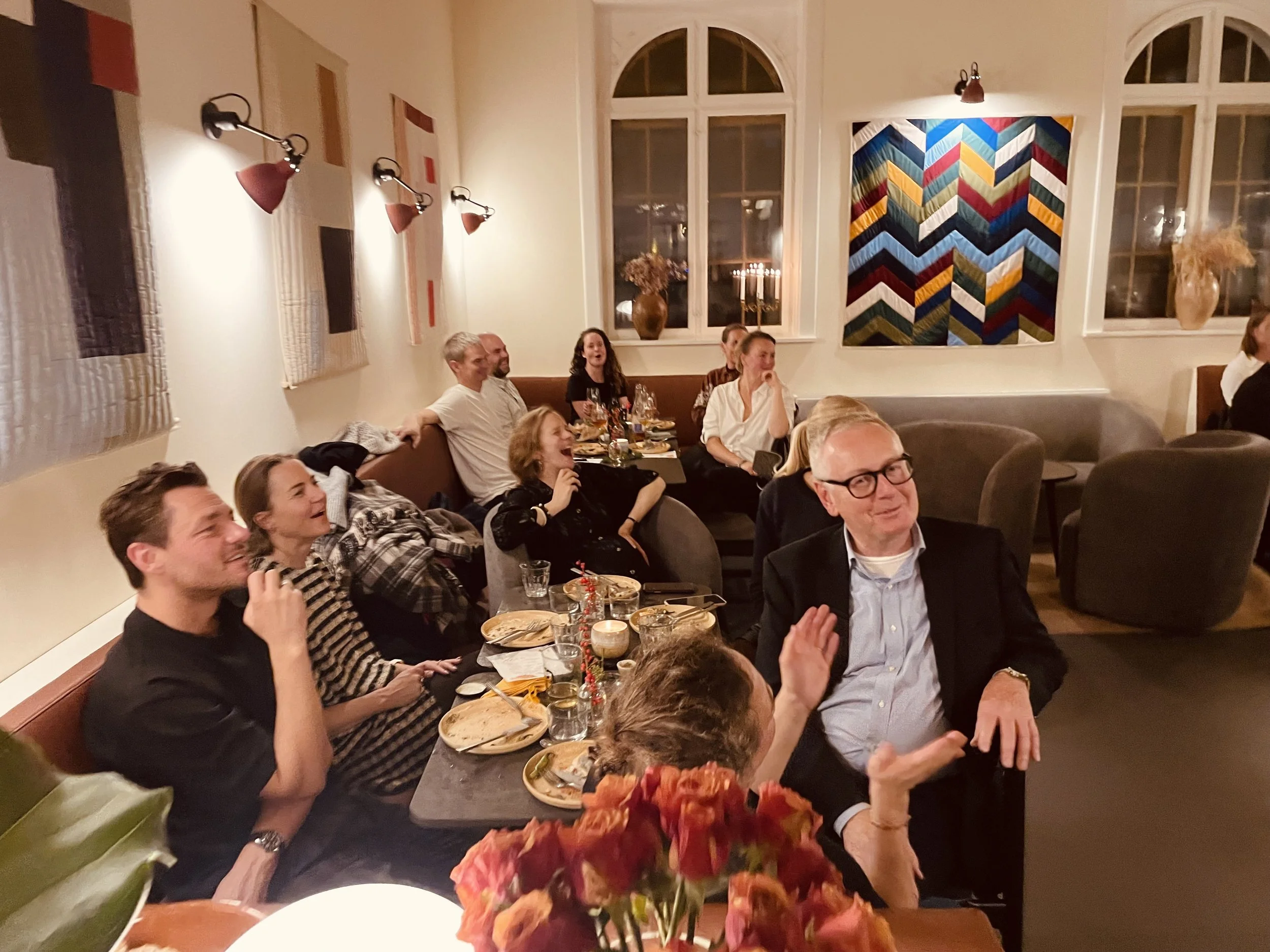 Gruppe mennesker sidder ved borde i en hyggelig stue, de er glade og griner, med mad og drikke på bordet, i baggrunden er der store vinduer og moderne vægdekoration.