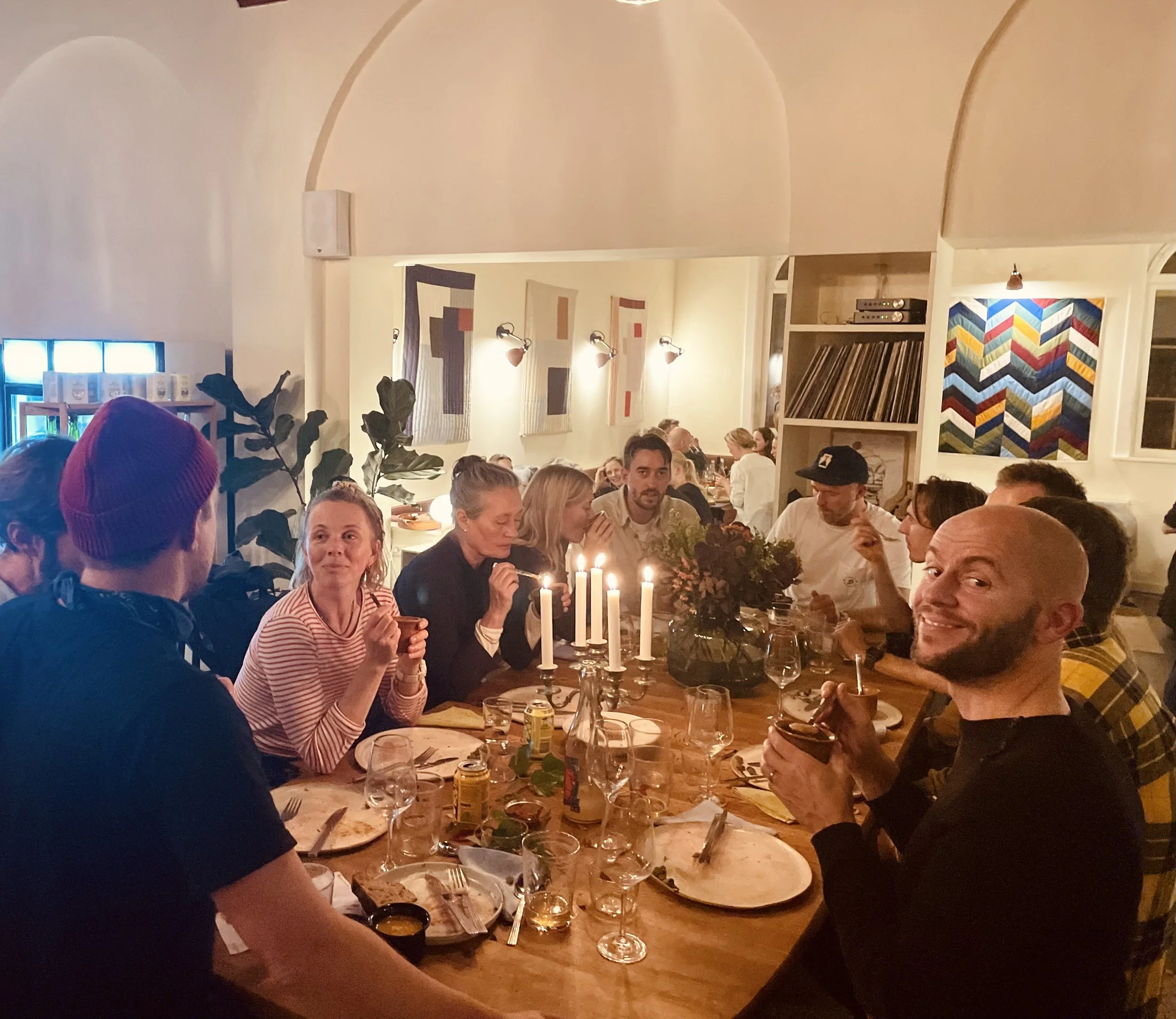 Gruppesamling af mennesker til middag, dekoreret med stearinlys og blomster på et bord i et hyggeligt indendørs rum.