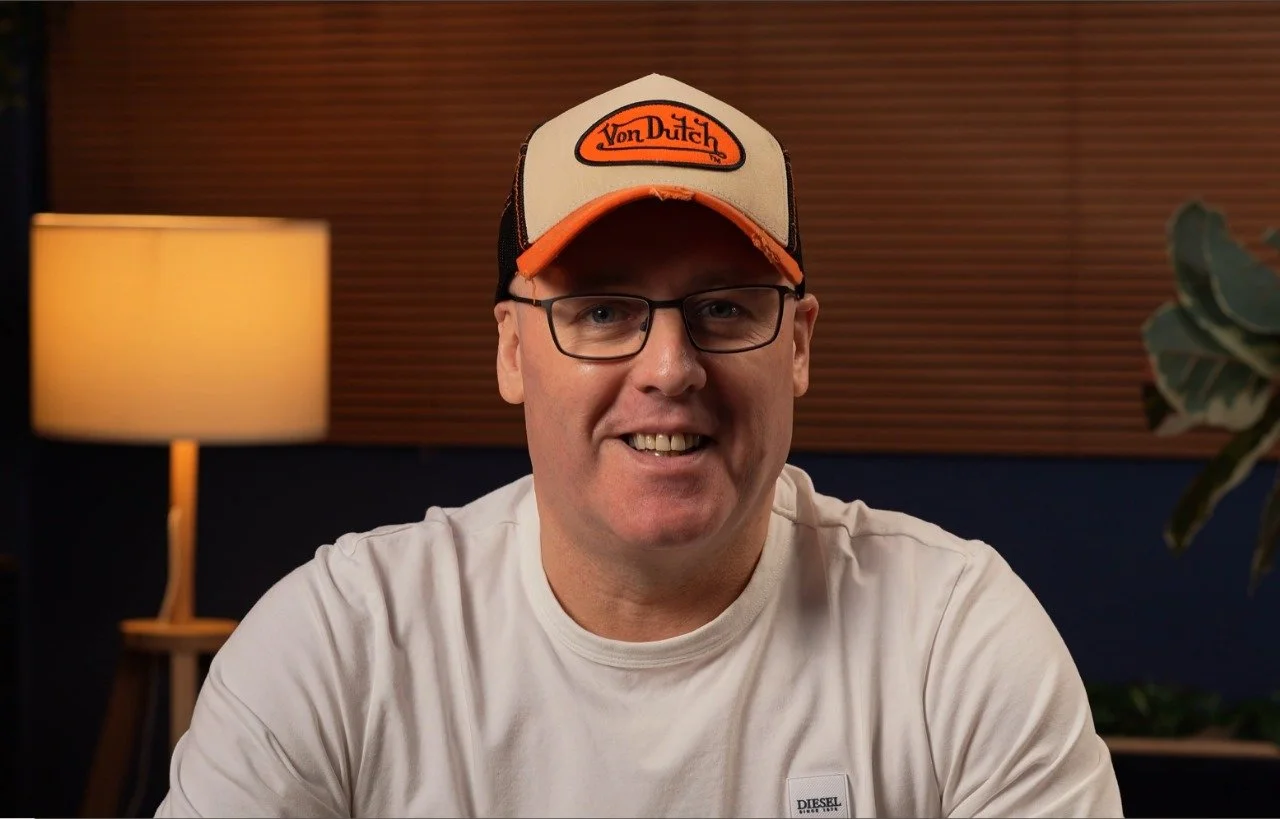 A man wearing glasses, a beige and orange Von Dutch trucker hat, and a white t-shirt sitting indoors near a lamp and a plant.