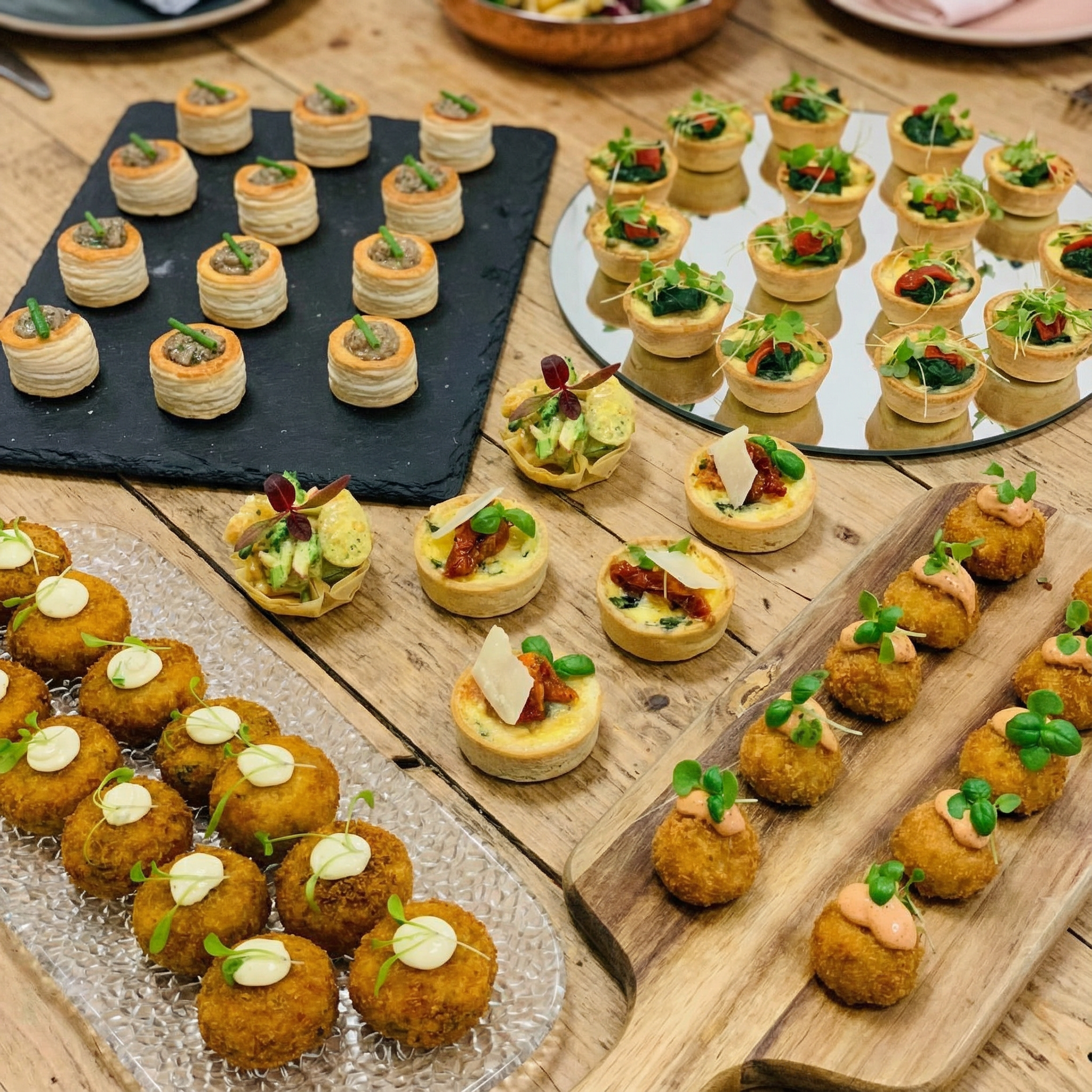 Plateaux de petites bouchées apéritives variées, dont des rouleaux de pâte feuilletée, des tartlets aux légumes, et des croquettes frites décorées de micro-pousses, sur une table en bois.