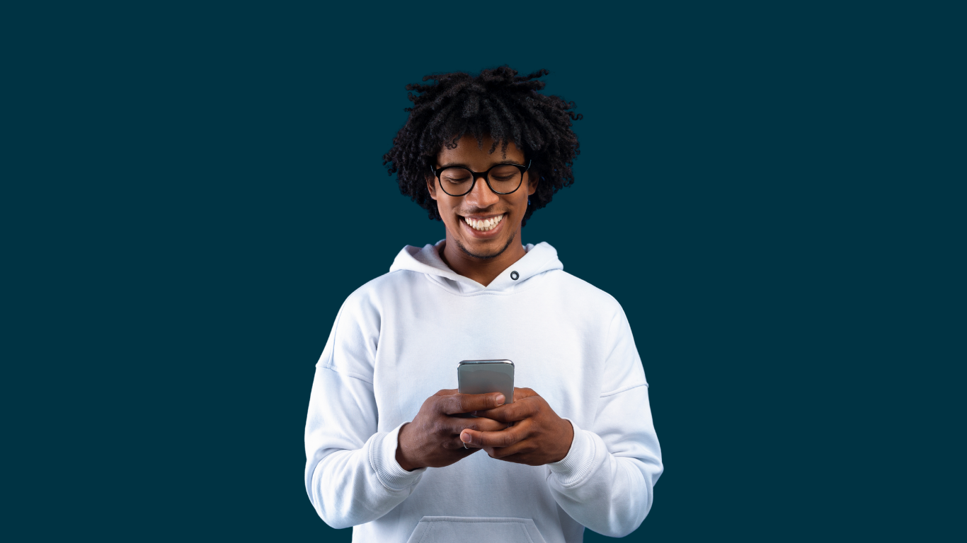 Young man with glasses smiling while looking at his phone, wearing a white hoodie, against a dark teal background.