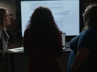 Three women looking at a large screen displaying a presentation or document, in a dimly lit room.