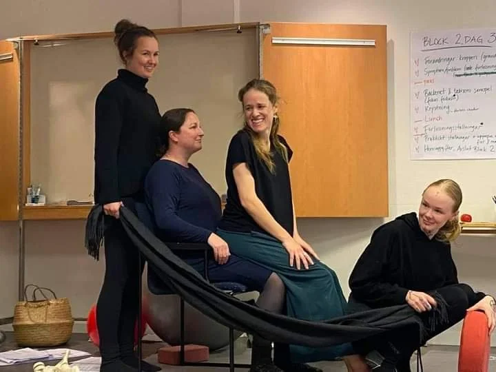 Four women in a classroom or office setting, with one standing and three sitting, interacting and smiling.