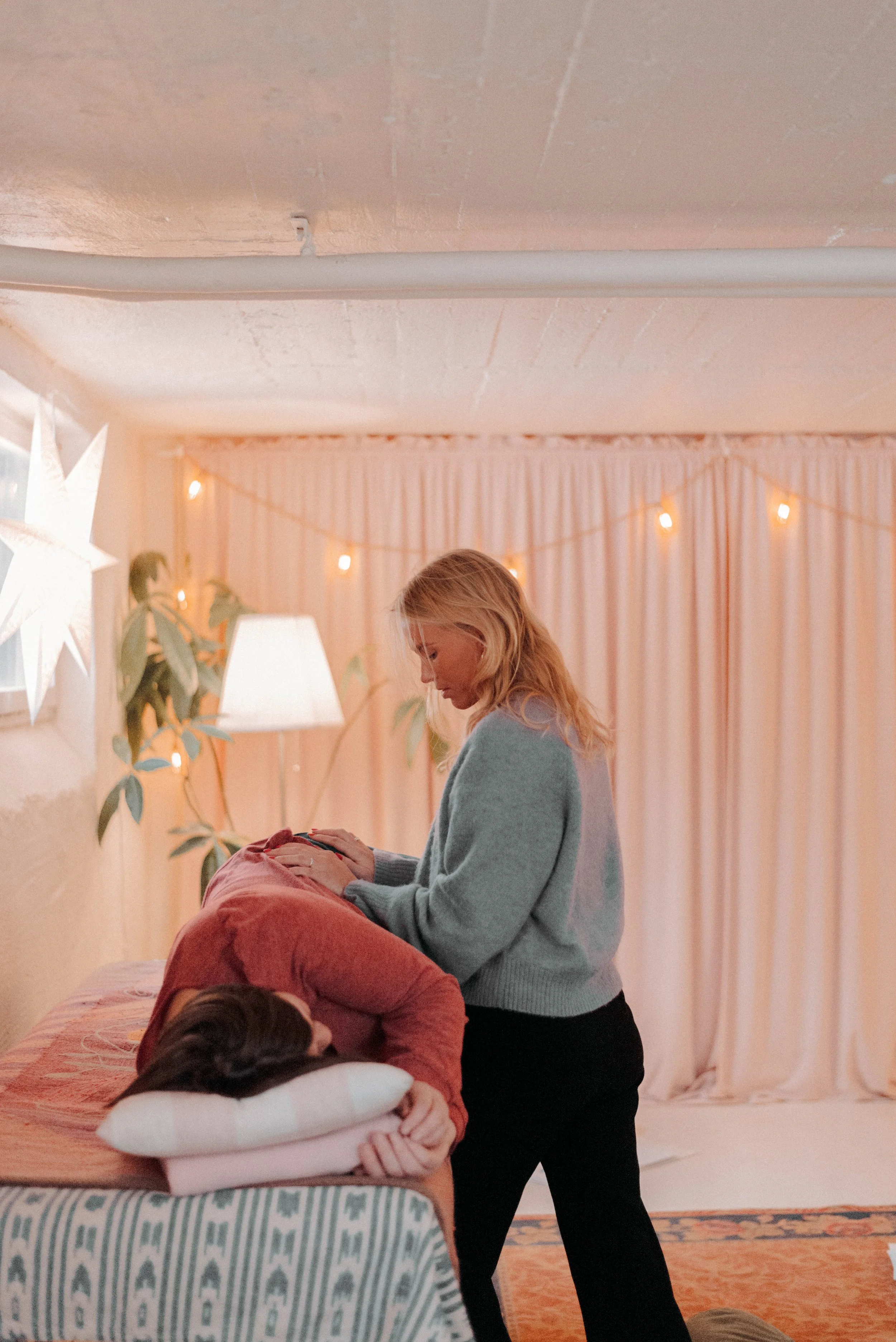 A woman attending a birth where a person is lying on a table, the woman is holding the person's hand while they are in labor. The setting is warm with soft lighting, pink curtains, decorative string lights, indoor plants, and stars on the wall.