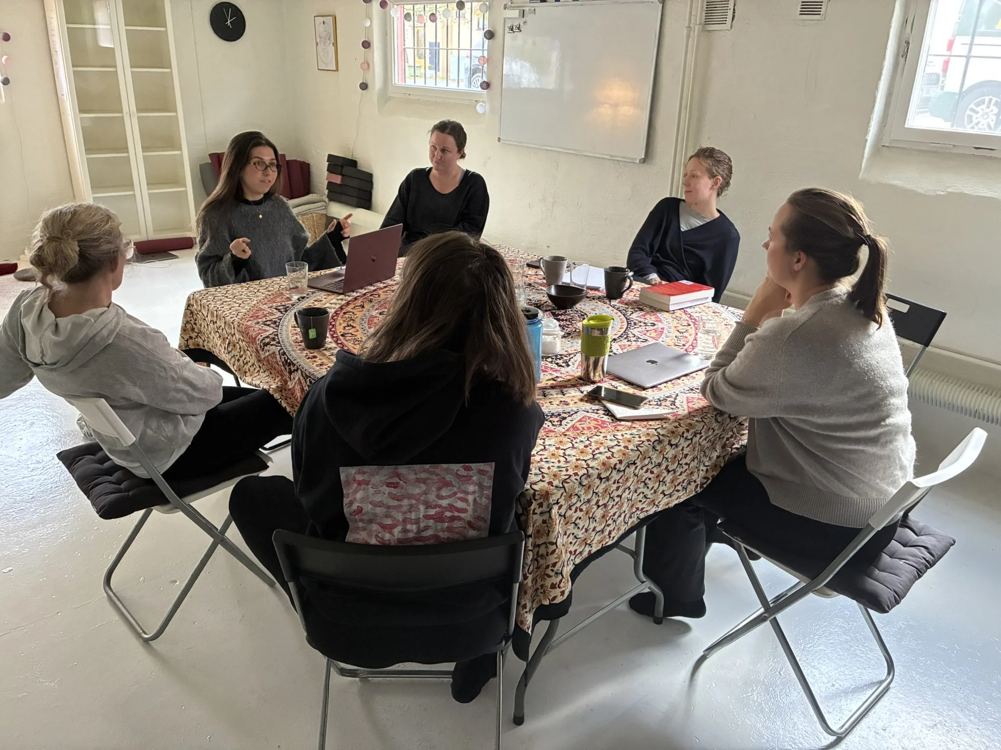 A group of six women sitting around a table in a bright room, engaged in a discussion. The table has various cups, a book, and a laptop. The room has light-colored walls, windows, and decorative string lights.