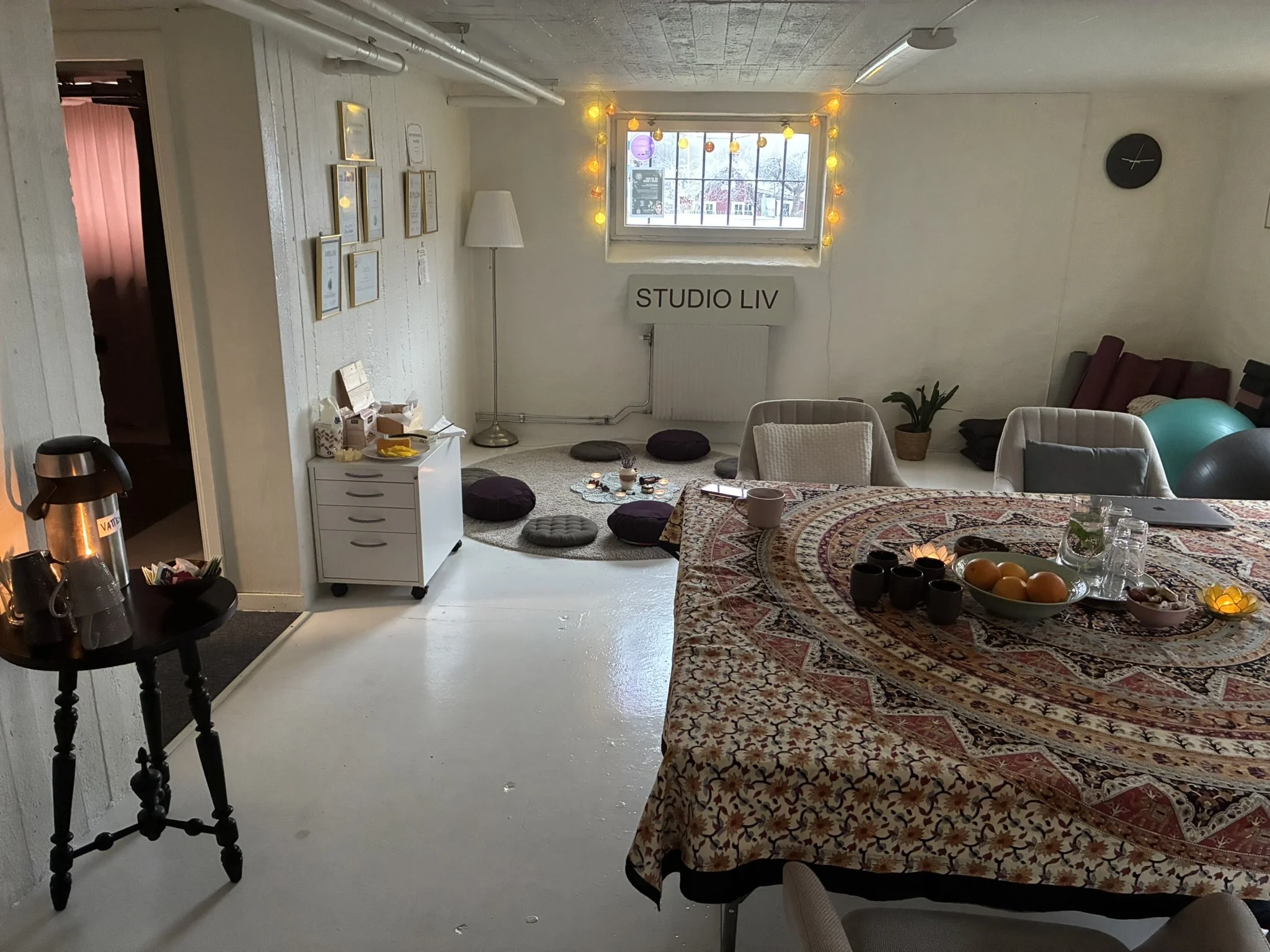 Cozy room with white walls, decorated with framed certificates, a lamp, and string lights around the window. A sign reads 'STUDIO LIV.' There are cushions on the floor, a table with a patterned tablecloth, and a fruit bowl, indicating a comfortable living or work space.