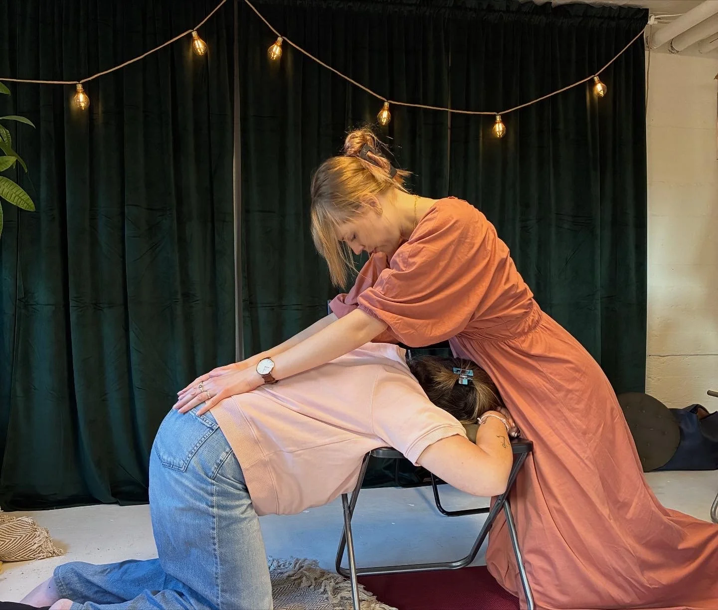 A woman receiving a massage from another woman in a room with dark green curtains and string lights.