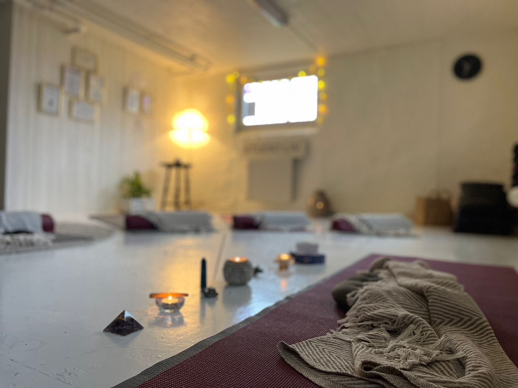 Low-angle view of a cozy living room with a lit lamp, framed pictures on the wall, and a window decorated with string lights. A rug with blankets and cushions is visible in the foreground.