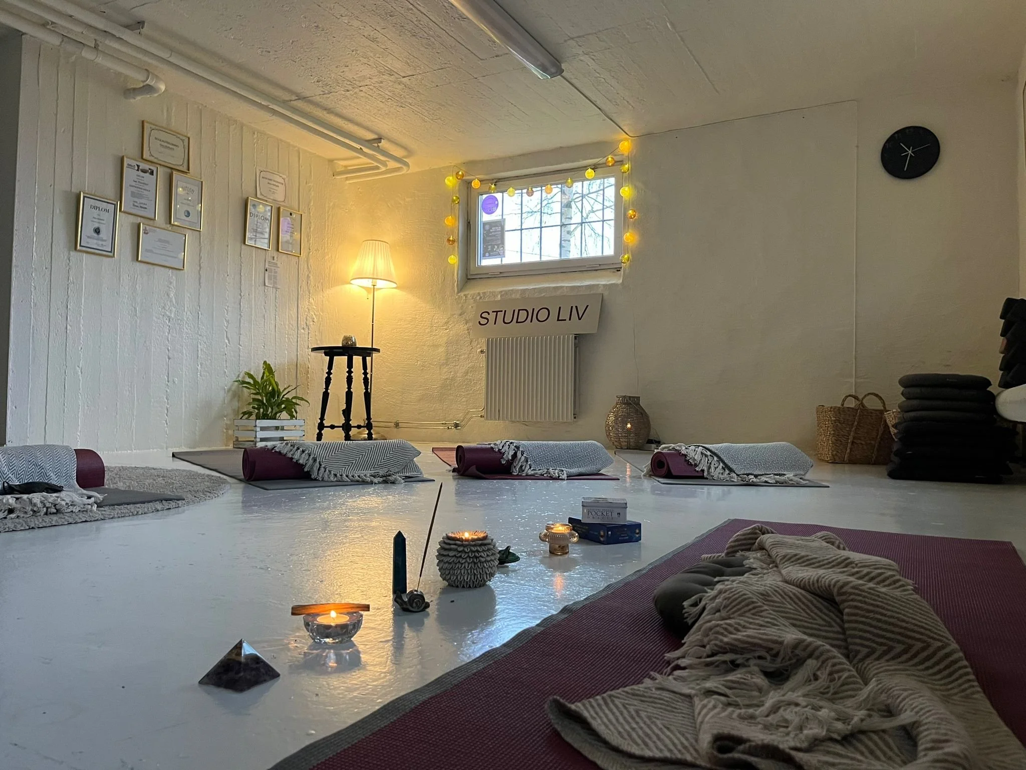 A cozy room with white painted brick walls decorated with framed certificates, a small window with fairy lights, a standing lamp, and yoga mats with cushions on the floor for a calming practice or meditation session.
