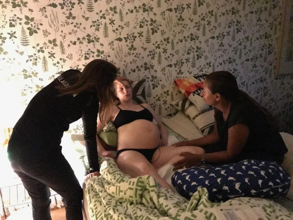 A woman lying in a bed with neck and abdomen injuries, being attended to by two women, one on each side, in a room with floral wallpaper.