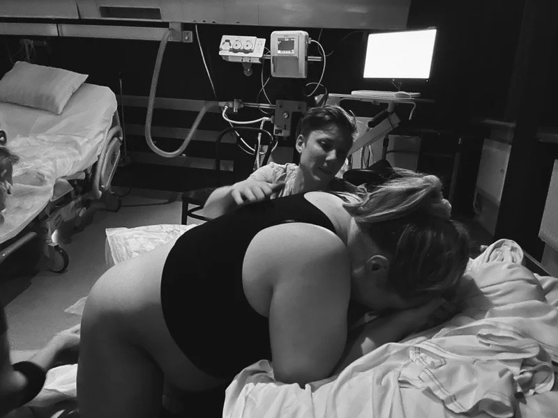 A woman laboring in a hospital bed with her head down, assisted during childbirth. A young man stands nearby, looking on, while a computer monitor and medical equipment are visible behind them.