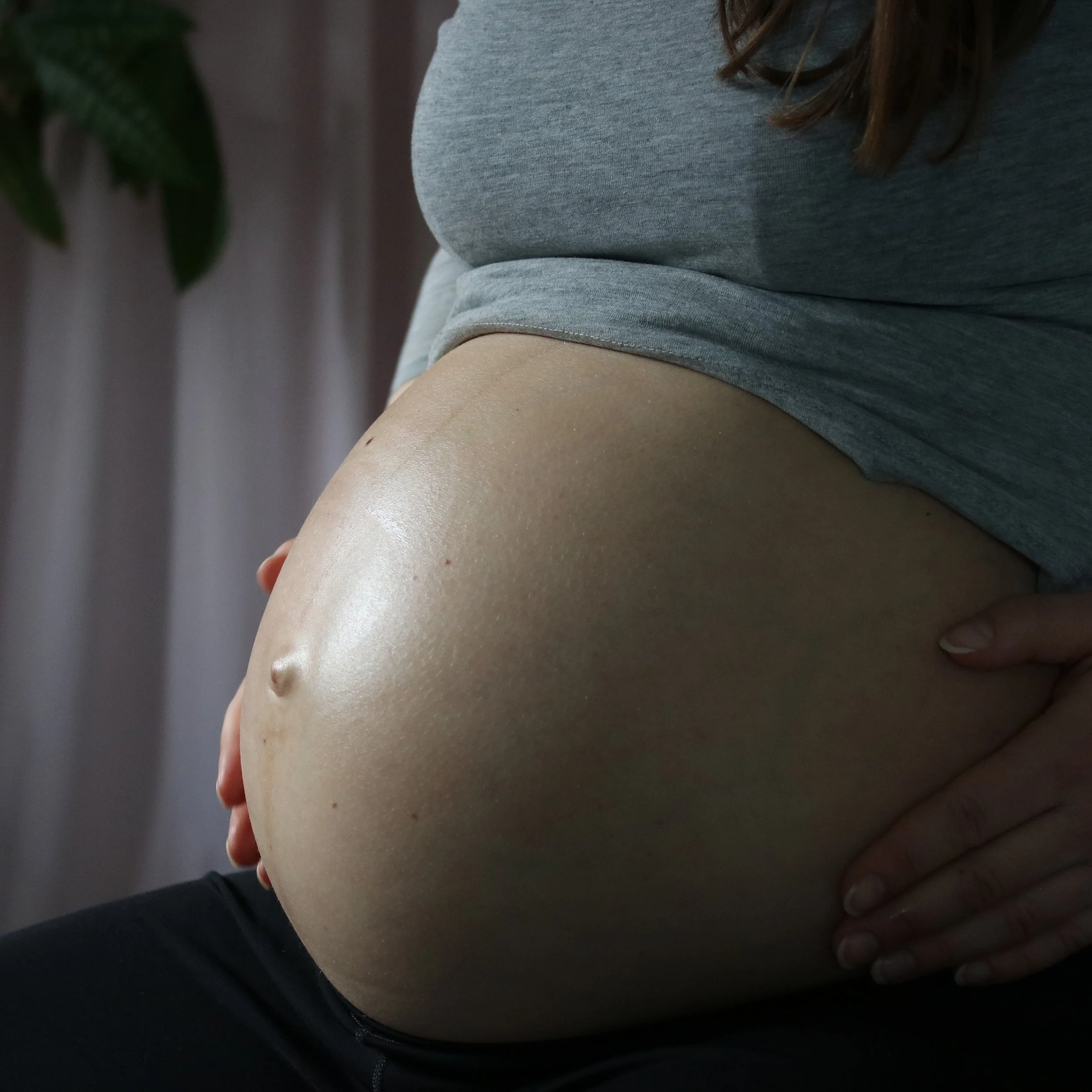 Close-up of a pregnant woman's belly, with a hand resting on her stomach, wearing a gray shirt.