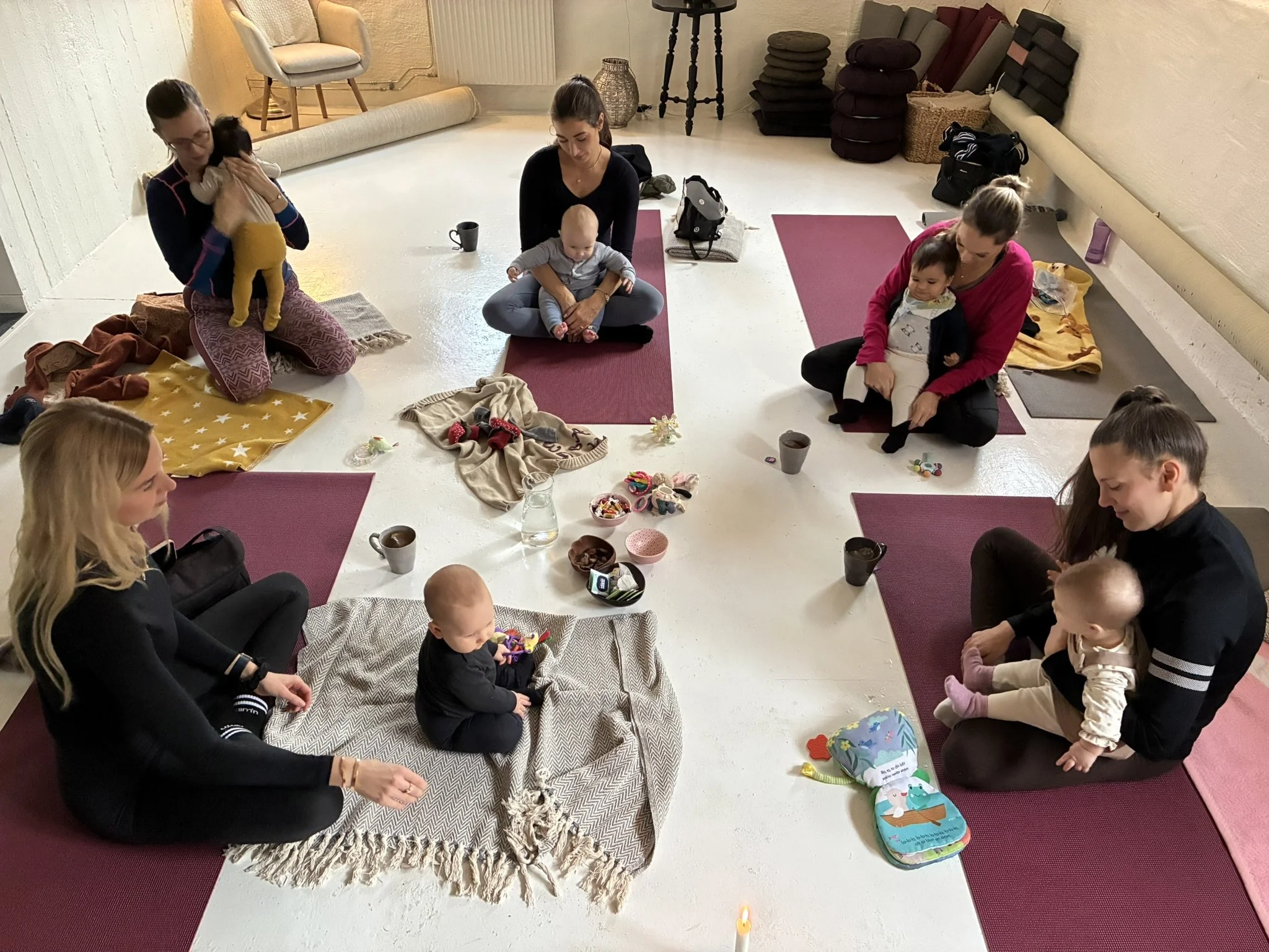 A group of women and children sitting on yoga mats in a circle, engaging in a group activity or meditation in a room with white walls, a rolled-up carpet, and various cushions and bags in the background.