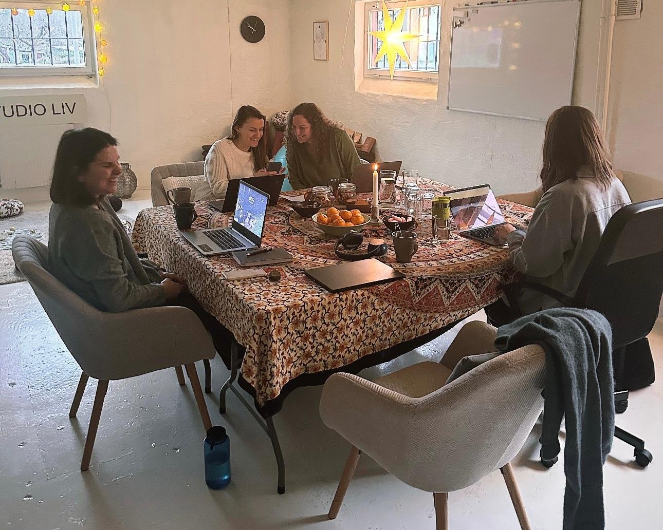 Five women gathered around a large, patterned table in a bright room, engaged in a meeting with laptops, notebooks, and snacks, with candles and warm lighting creating a cozy atmosphere.