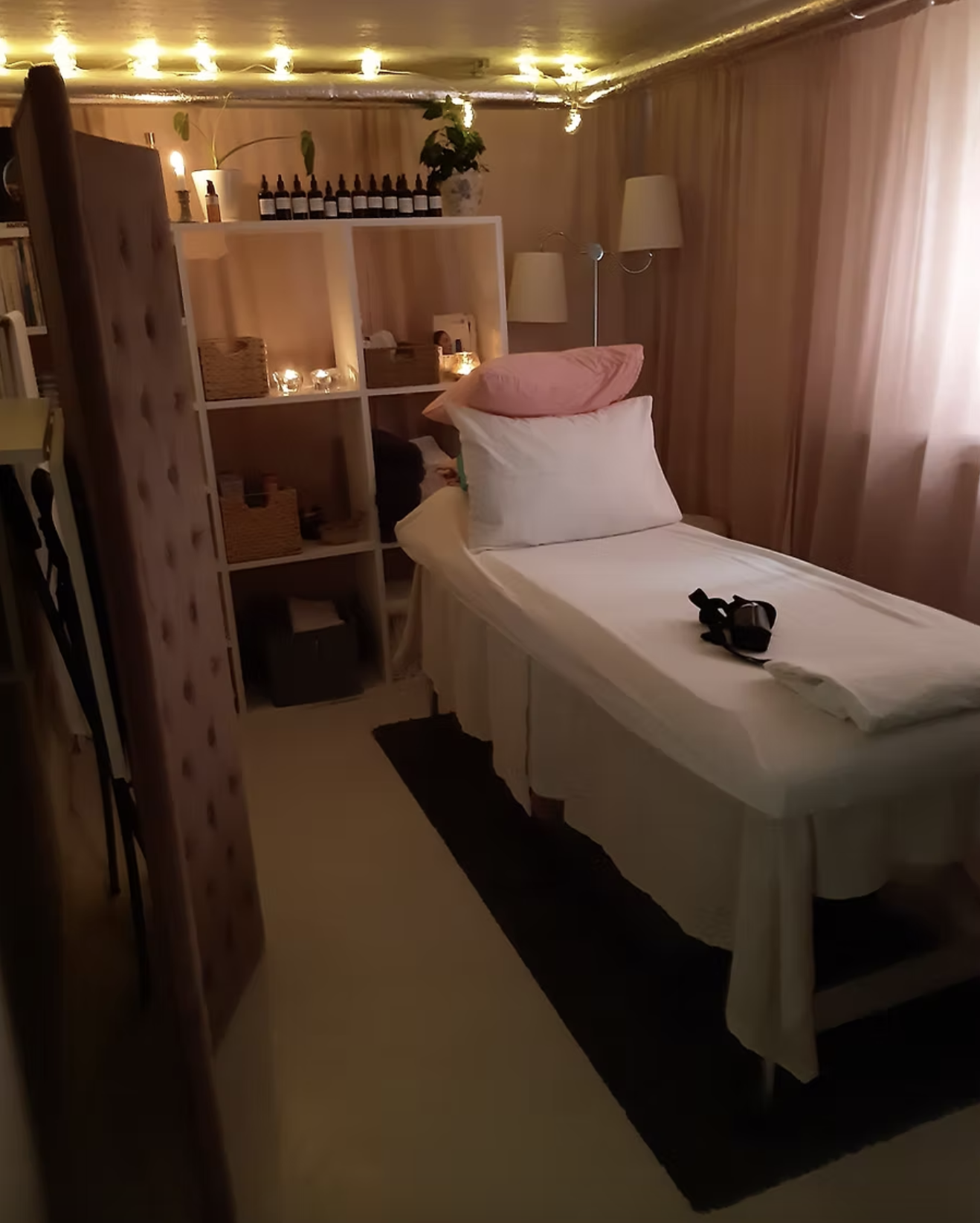 A cozy bedroom with soft pink curtains, a bed with white sheets, pillows, and a black headphone resting on the bed. There are lanterns and candles on shelves, a tall floor lamp, and string lights along the ceiling, creating a warm and inviting atmosphere.